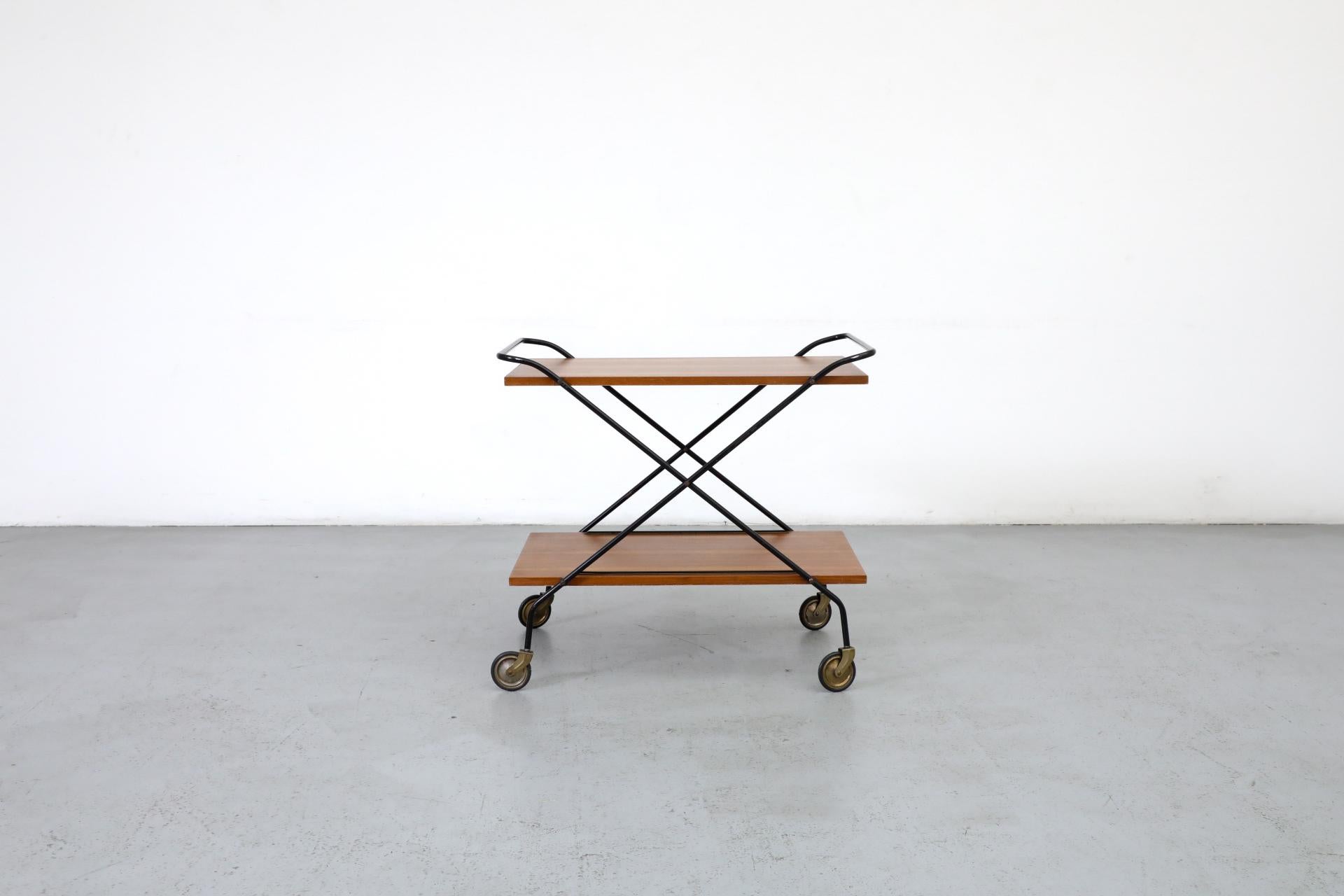Mid-Century two tiered rolling bar cart with black enameled metal frame and 2 teak shelves with wonderfully patterned grain by by Paul Nagel for Jie Gantofta, 1960s. Shelves are painted white on undersides. Larger brass wheels. Bottom shelf has