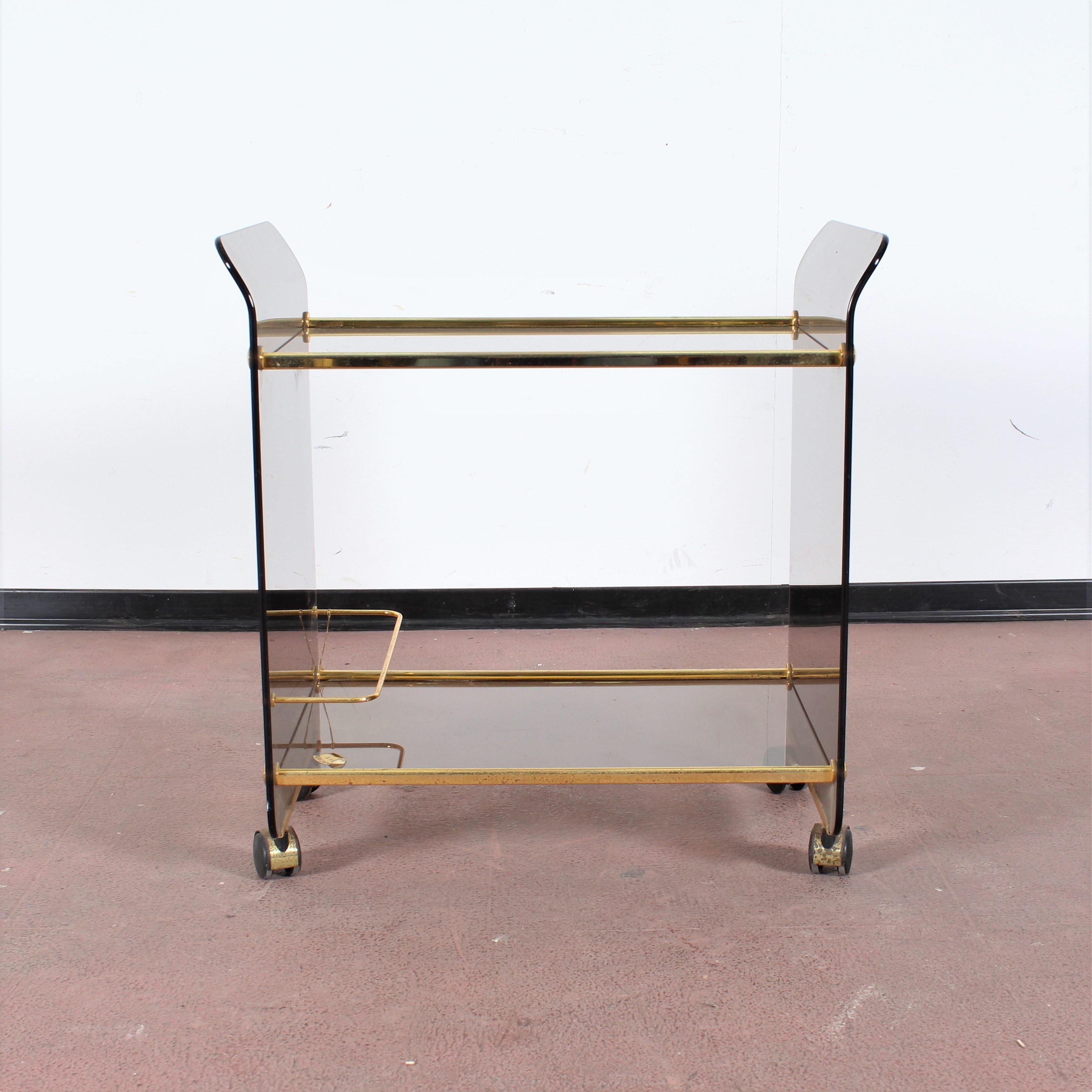 Beautiful two-tier bar cart on wheels, in curved smoked glass, with elements in 24-carat gold-plated brass. Design by Umberto Mascagni, 1960s, Italy.
The original brand on the glued label can be read on the underlying surface, slightly worn on the