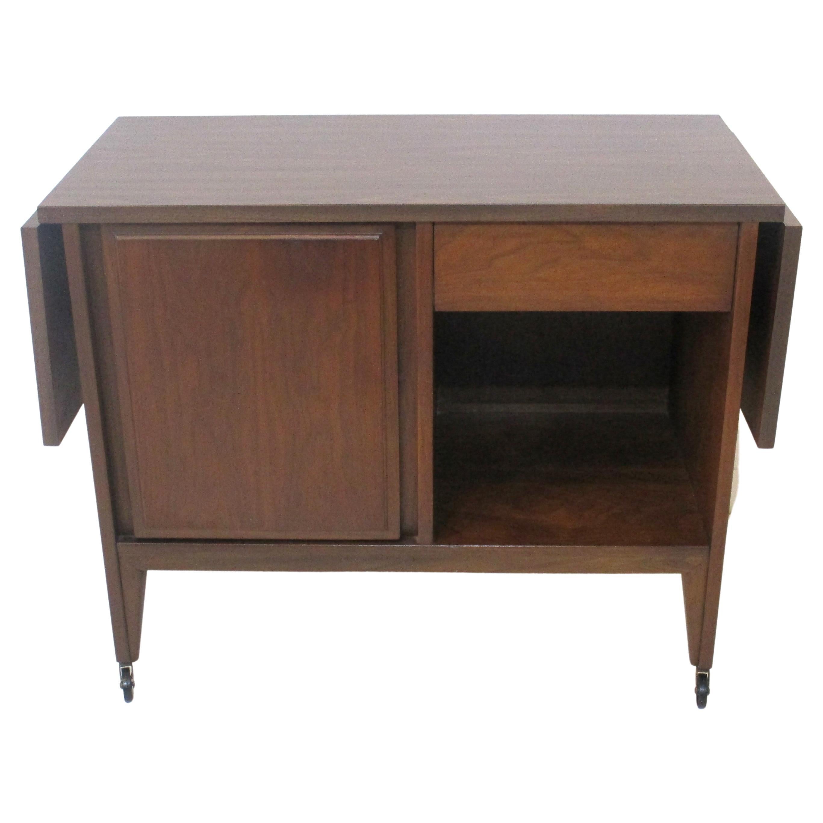 A medium walnut rolling bar cart with folding formica covered top making for a larger mixing and party area. Below there's a door with storage for bottles and glasses with a shelve, a upper drawer for bar tools and an additional open storage area.