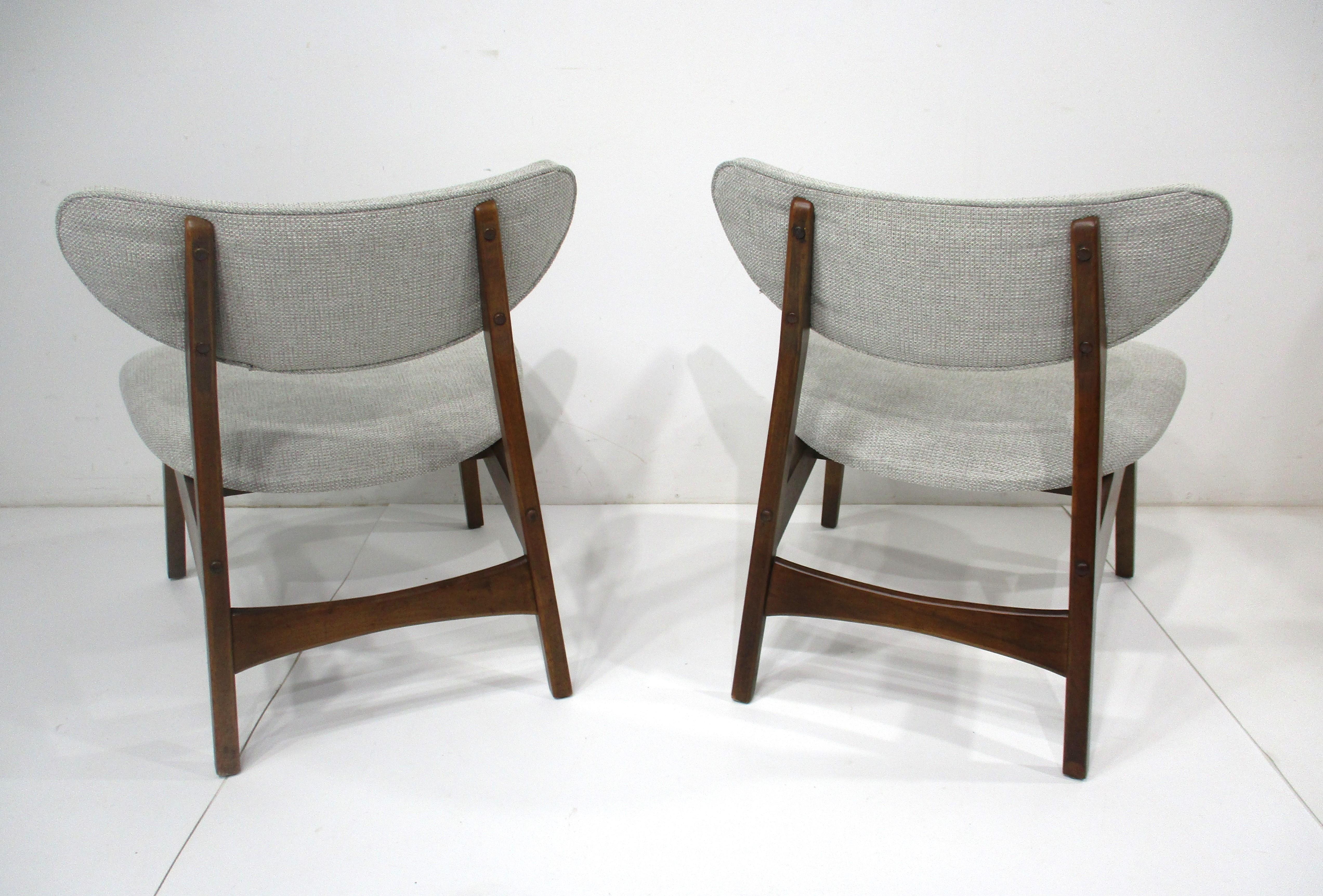 A pair of low lounge chairs with dark walnut toned wood frames and upholstered bottom and curved back . Having a very comfortable sitting position with plenty of support for long periods of relaxing . Designed in the manner of Danish Modern and