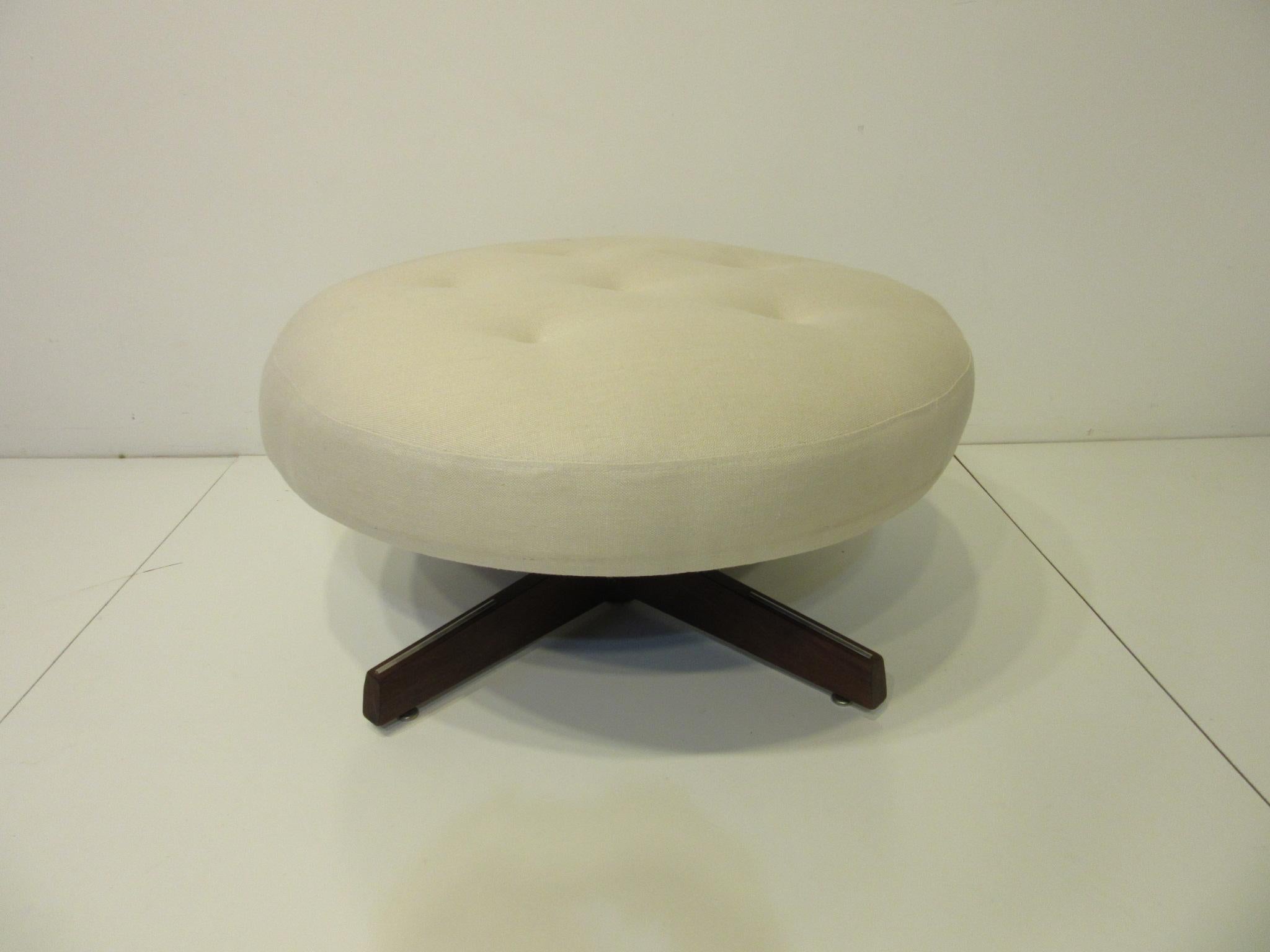 A well crafted upholstered round swiveling ottoman in a cream colored Italian styled linen with button cushion top, walnut four star base detailed with brushed metal strips and having adjustable foot pads. Can be used as extra seating, for a lounge