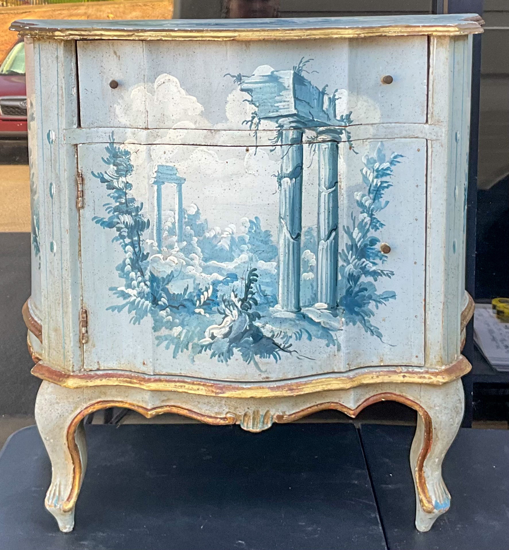 This is a lovely little Venetian commode. It is hand painted with Italian Neo-classical scenes in soft blues and gilt. It is a single drawer over open door. Age wear throughout the piece. It most likely dates to the 50s.
