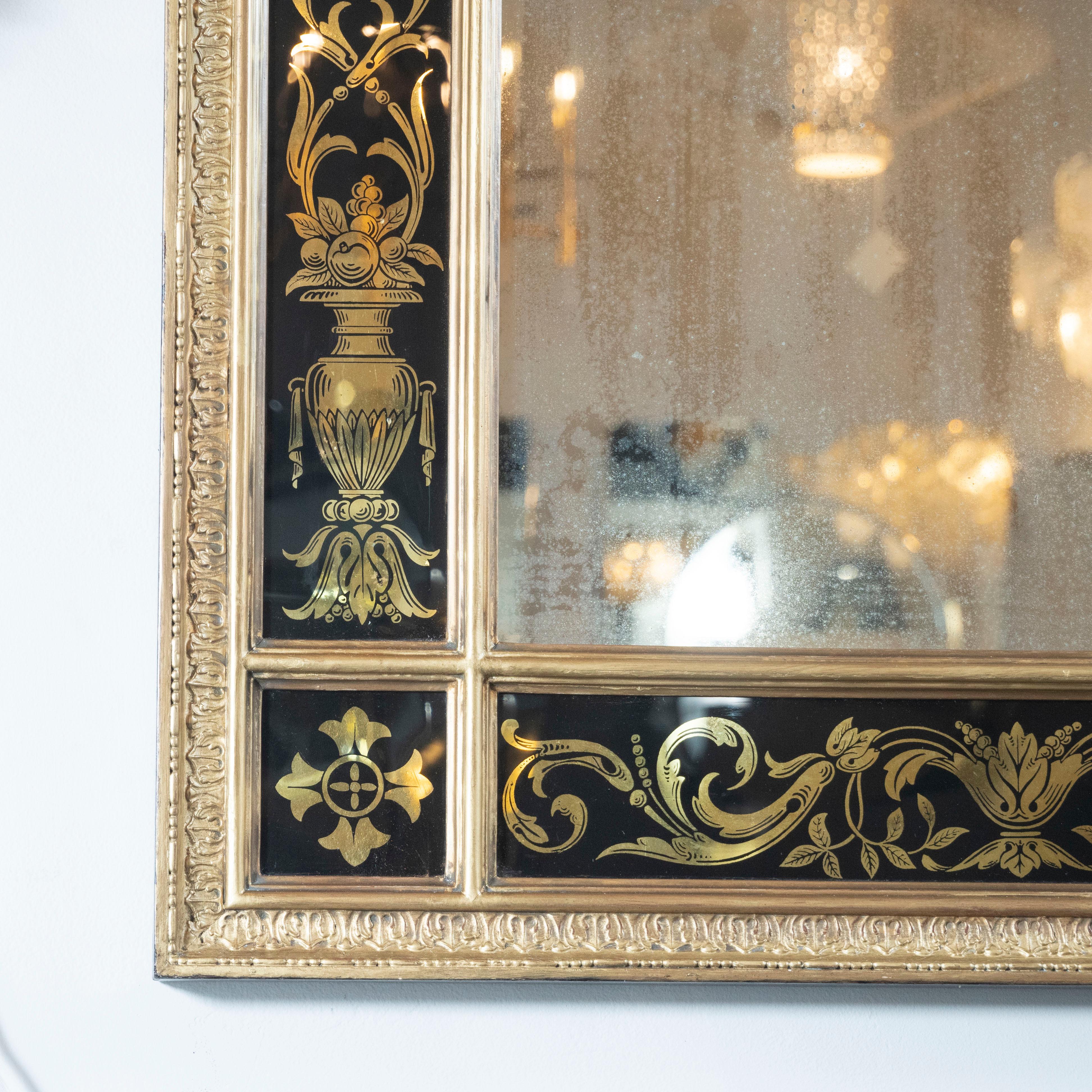 This elegant Mid-Century Modern Venetian wall mirror was realized in Italy, circa 1950. It features a filigreed gilded frame with reverse églomisé offering foliate and figurative motifs against a black background with rectangular plain mirror in the