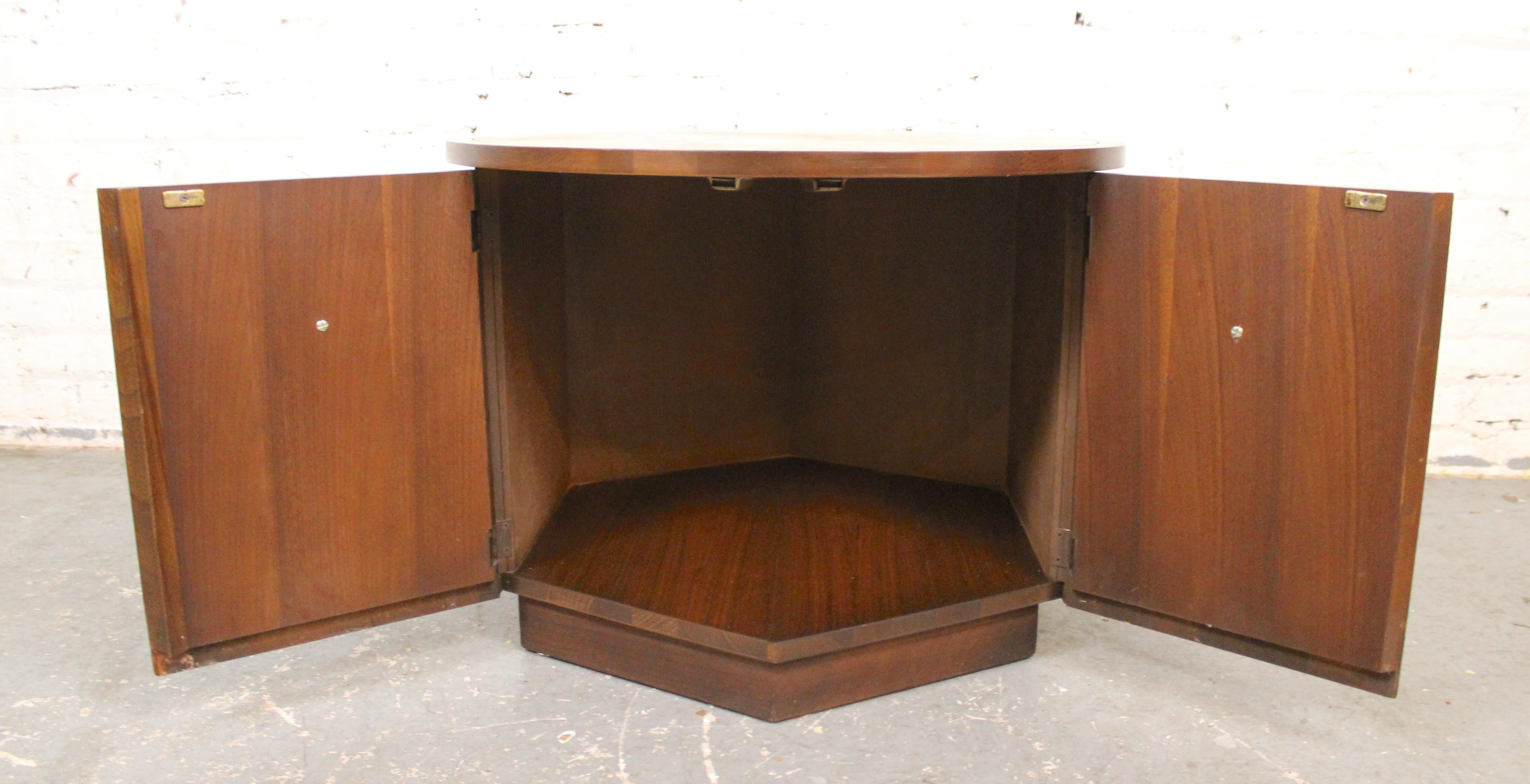 Runder Beistelltisch aus amerikanischer Produktion (wahrscheinlich von Bassett Furniture) mit einem sechseckigen Sockel. Glamouröse Nussbaumfurniere zeigen die Maserung durchgehend, mit eingelegten Messinggriffen, die Doppeltüren für die