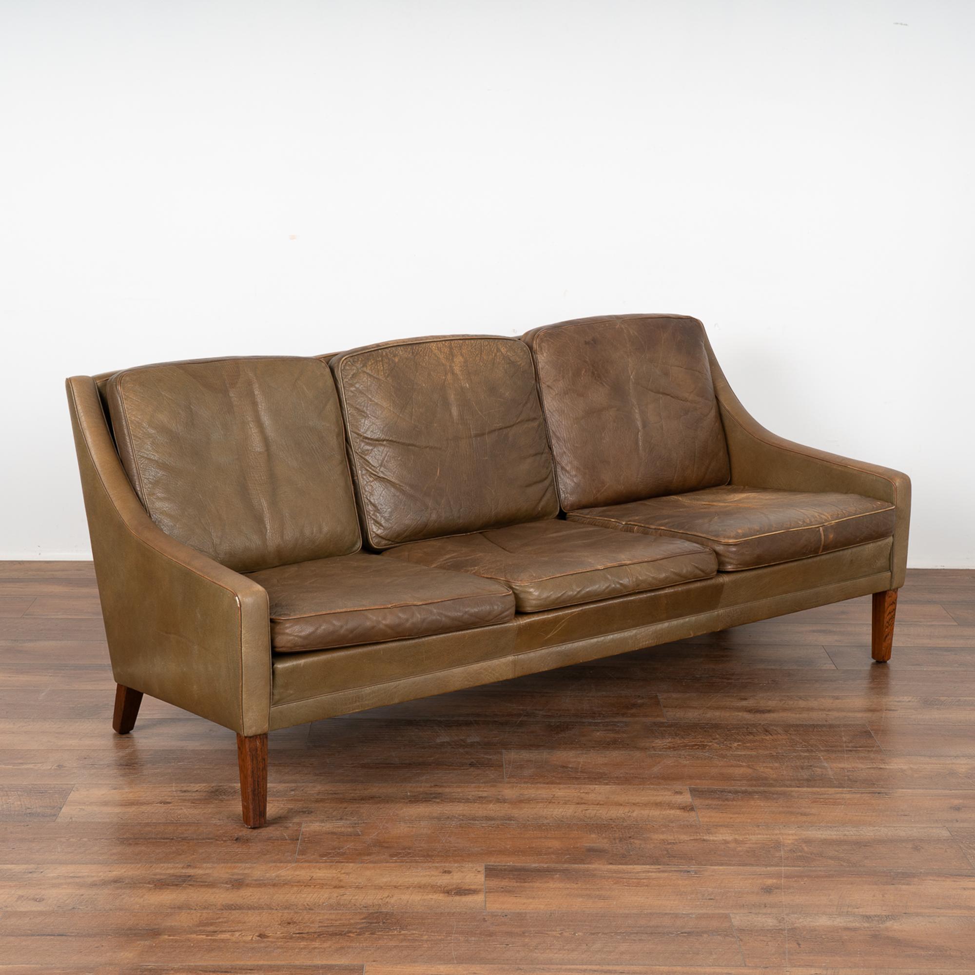 Mid-century modern brown leather three-seat sofa with removable cushions and hard wood frame.
The years of use are revealed in the aged patina of the leather, including impressions, scuffs/scratches, some discoloration, stains on seat cushions,