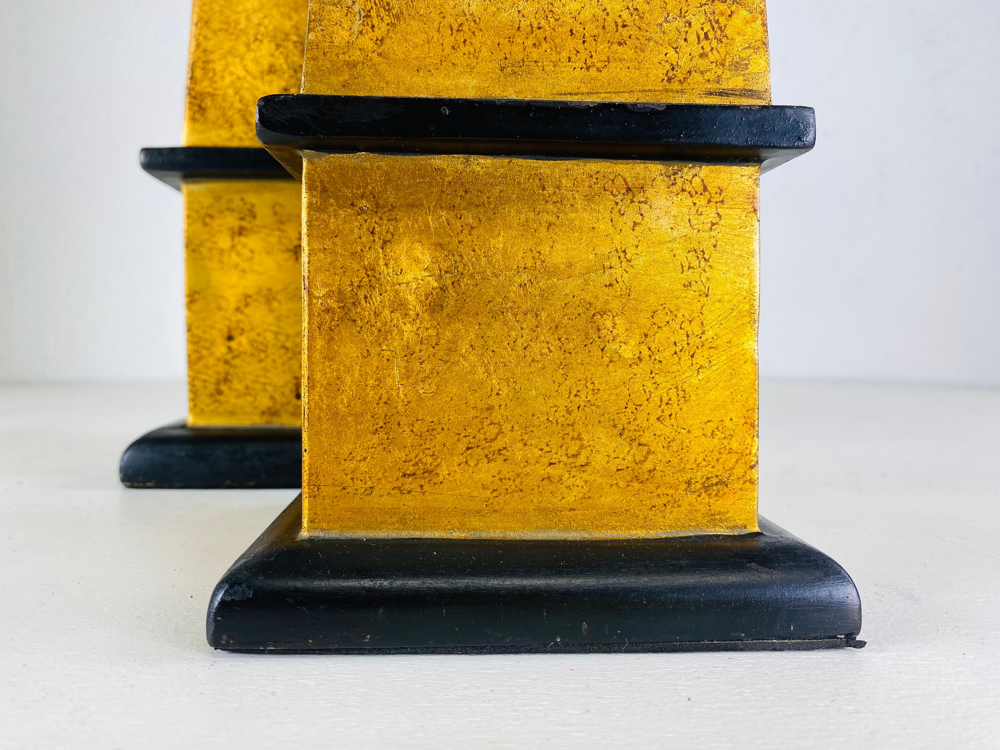 This is a pair of mid-century vintage wooden obelisks. These obelisks have a patinated bronze finish to the surface with a black trimmed detail at the base. The taller obelisk is 6 x 6 x 24. The smaller obelisk is 5 x 5 x 20.