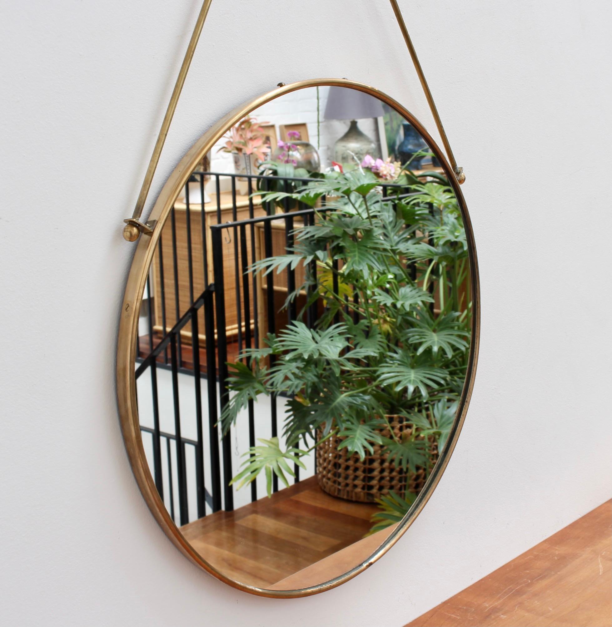 Mid-century Italian brass-framed wall mirror with decorative hanging rods (circa 1950s). The mirror is round with a characterful brass frame and unique hanging rods topped with a decorative finial. It is distinctive in the Italian modern style. The