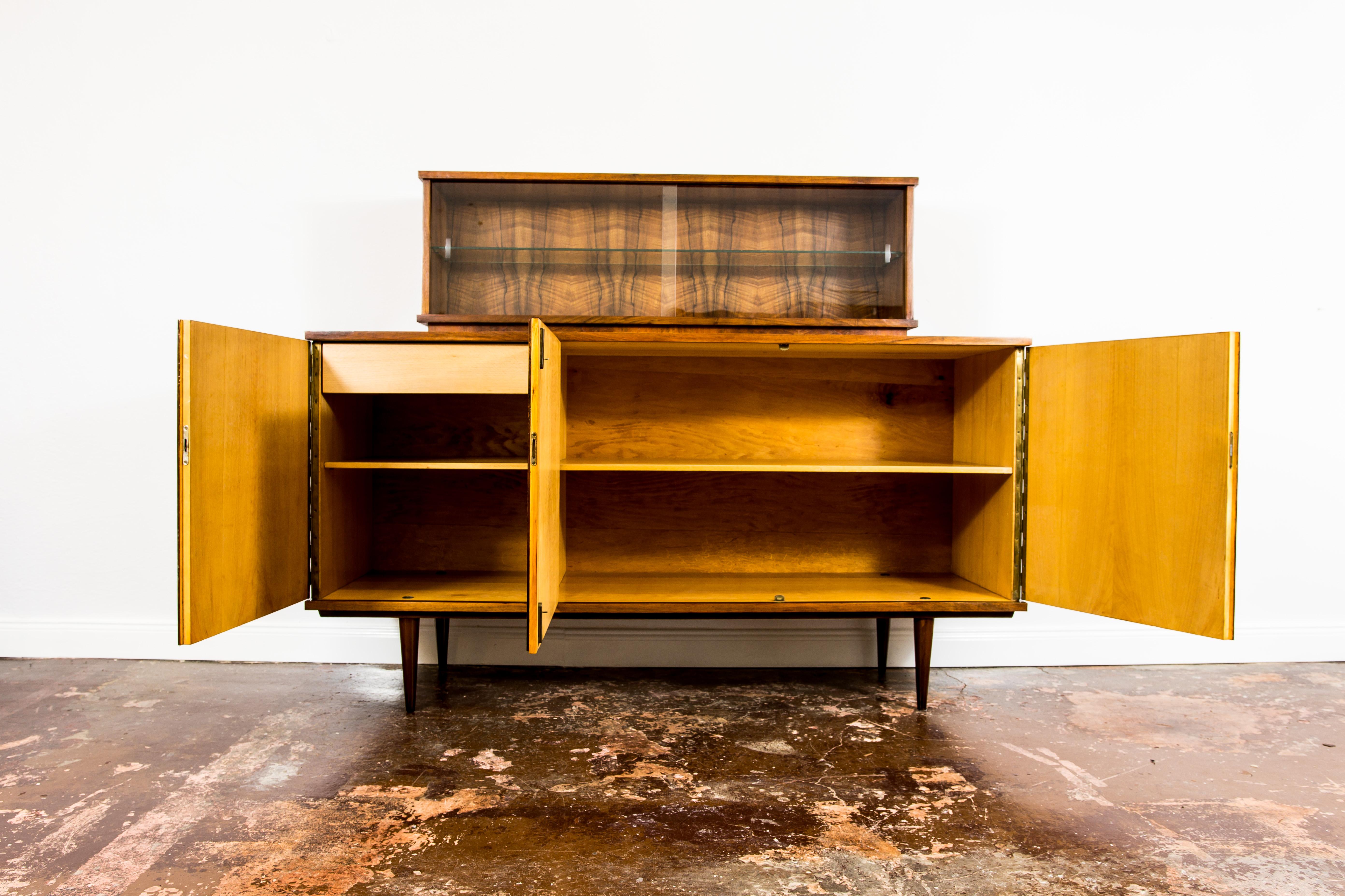 Vintage walnut veneer sideboard from Lódzkie Furniture Factories, made in Poland in 1960's 
Credenza has three doors, shelves and one drawer. This piece offers a display cabinet on the top comes with two clear glass sliding doors, and glass