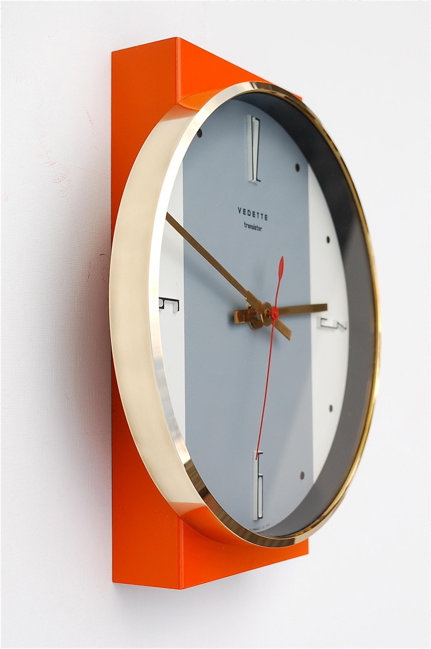 1960s vintage wall clock by French manufacturer Vedette. The circular clock face sits on a raised, orange lacquered, rectangular base. A broad grey stripe that runs across the face of the clock accentuates the vertical line of the design. The clock