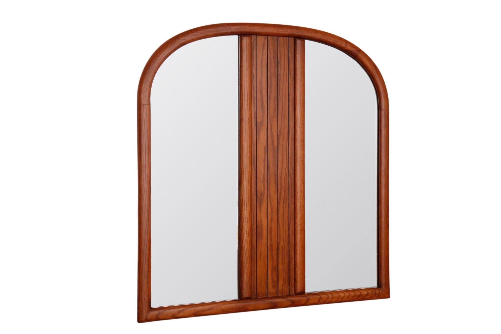 A mid century arched wall mirror made by Stanley Furniture. A wooden frame in oak is richly grained with a central beveled panel.