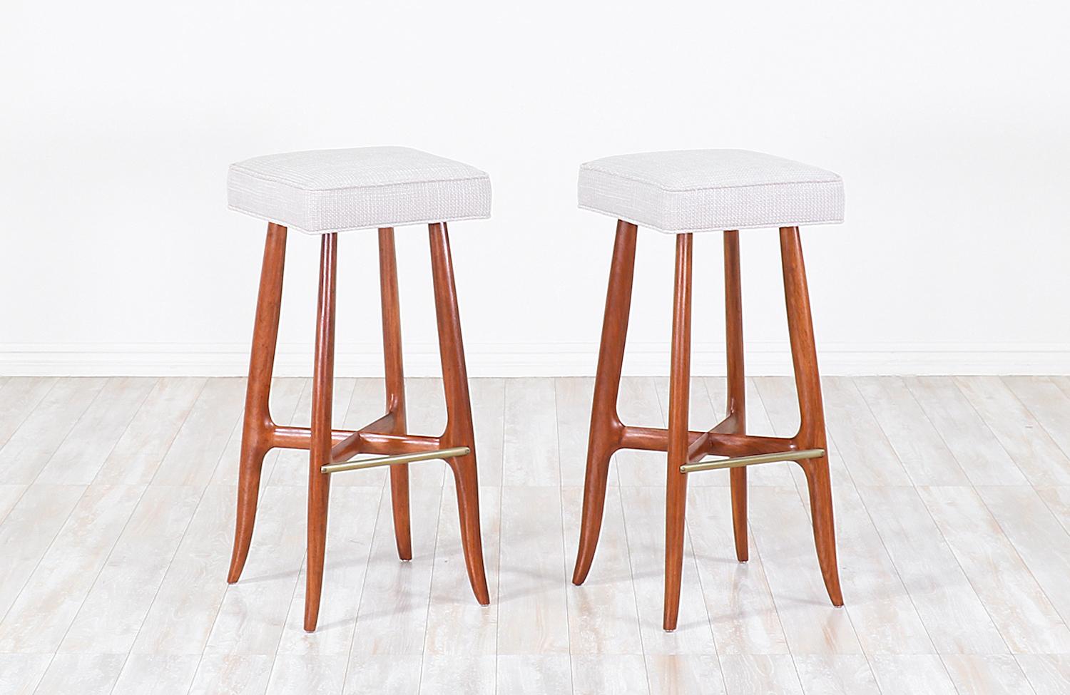 Pair of Mid-Century Modern bar stools designed and manufactured in the United States, circa 1960s. This Minimalist design features a walnut wood base with sculpted feet that flare out at the bottom, supporting wooden stretchers in the form of an X.