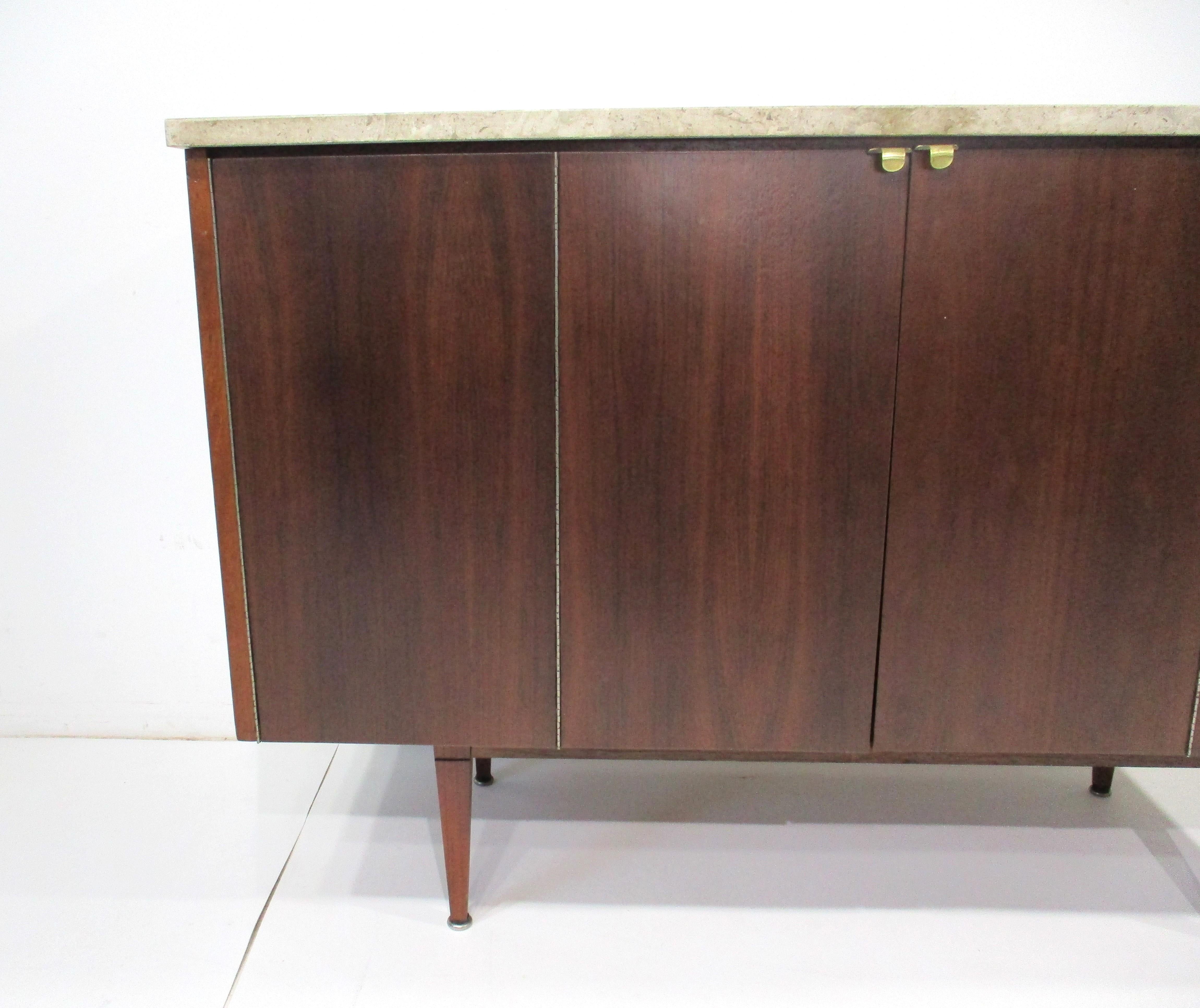 A Mid Century dark walnut bi -fold door bar cabinet with brass pulls sitting on tapered walnut legs . The top is in a medium tan / gray polished fossil marble making for a great conversational piece with outlines of prehistoric marine life over 65