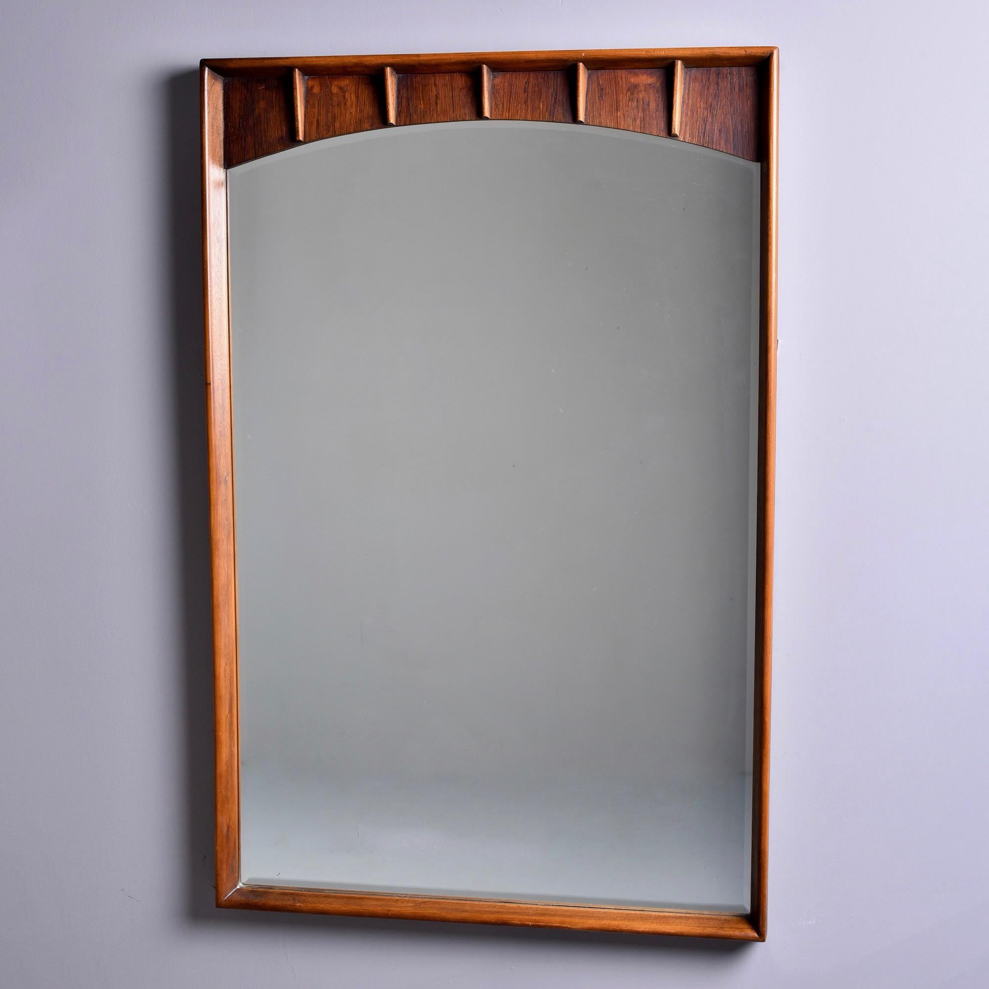Le miroir datant des années 1960 présente un subtil arc supérieur et un cadre profond en noyer et en pécan serti avec des accents étroits et nervurés au sommet. Les bords des miroirs sont biseautés. Trouvé aux États-Unis. Fabricant inconnu.