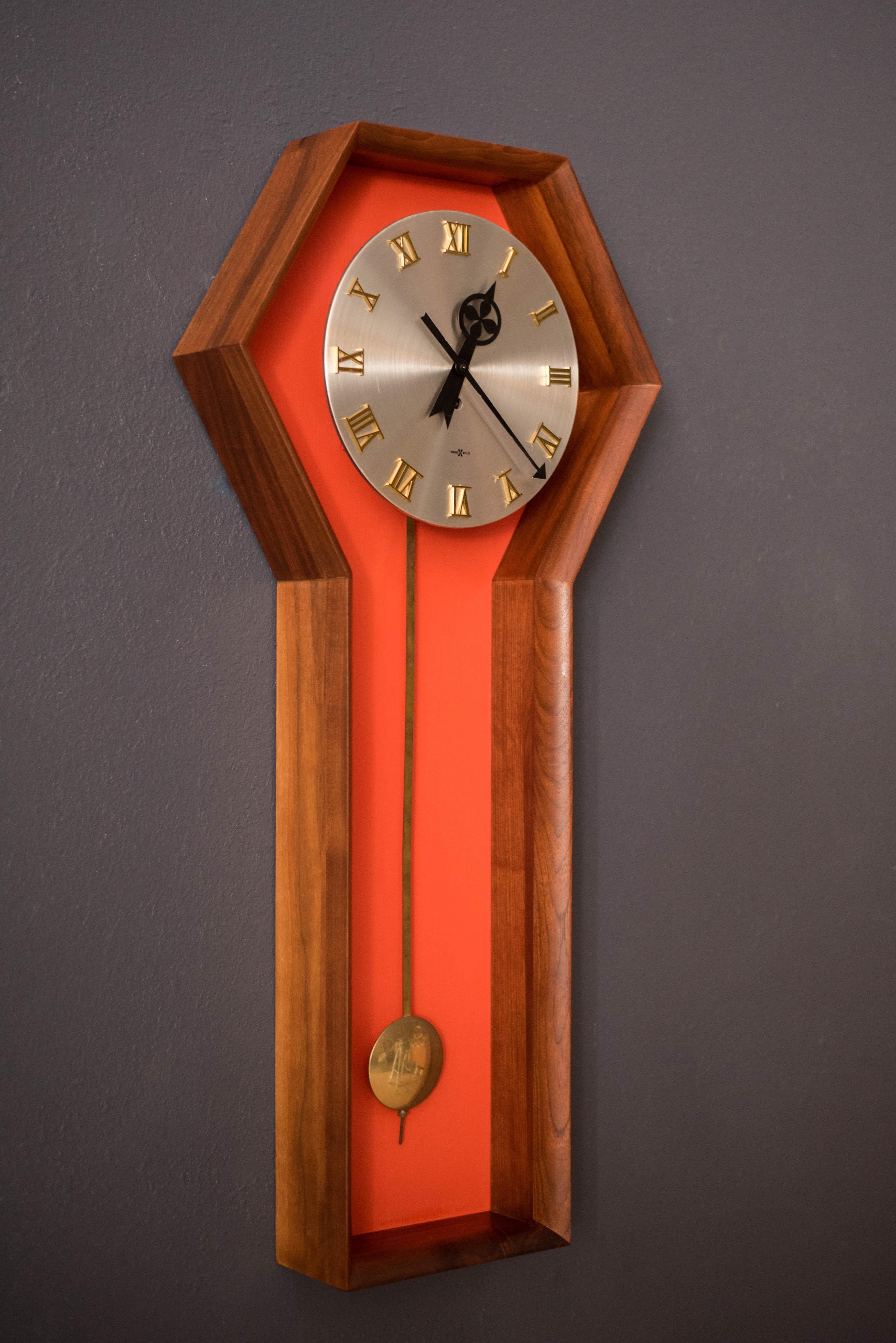 Vintage Meridian wall clock designed by Arthur Umanoff for Howard Miller in walnut, circa 1970s. This piece features a coffin shaped wood case and an inset orange backdrop accented with a brass pendulum. Includes the original clock hands and keeps