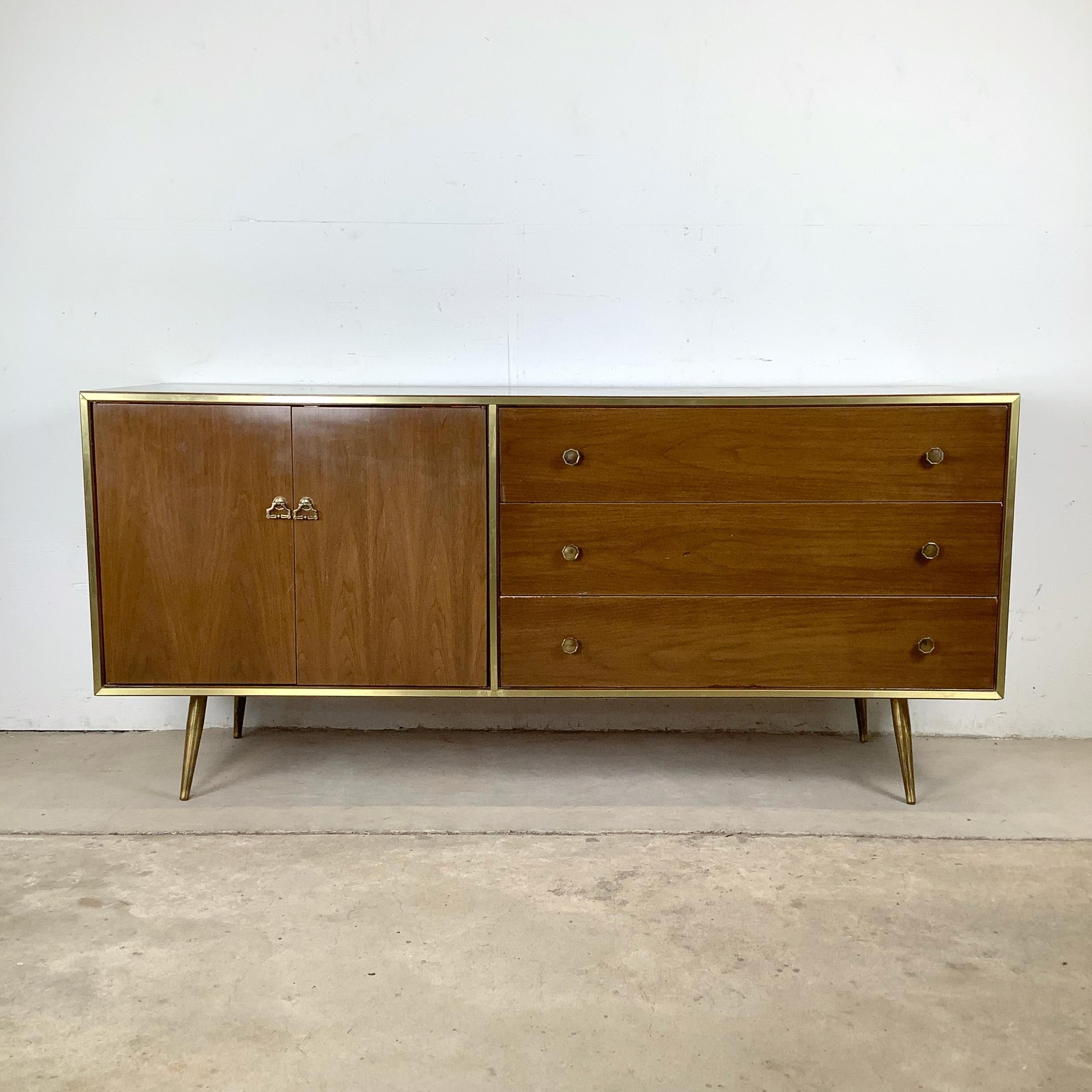 Questa bella cassettiera di metà secolo presenta un sorprendente equilibrio tra linee moderne e pulite e dettagli glamour. La cassa è rifinita con una calda impiallacciatura di noce incorniciata da finiture in ottone, le cui venature sottili