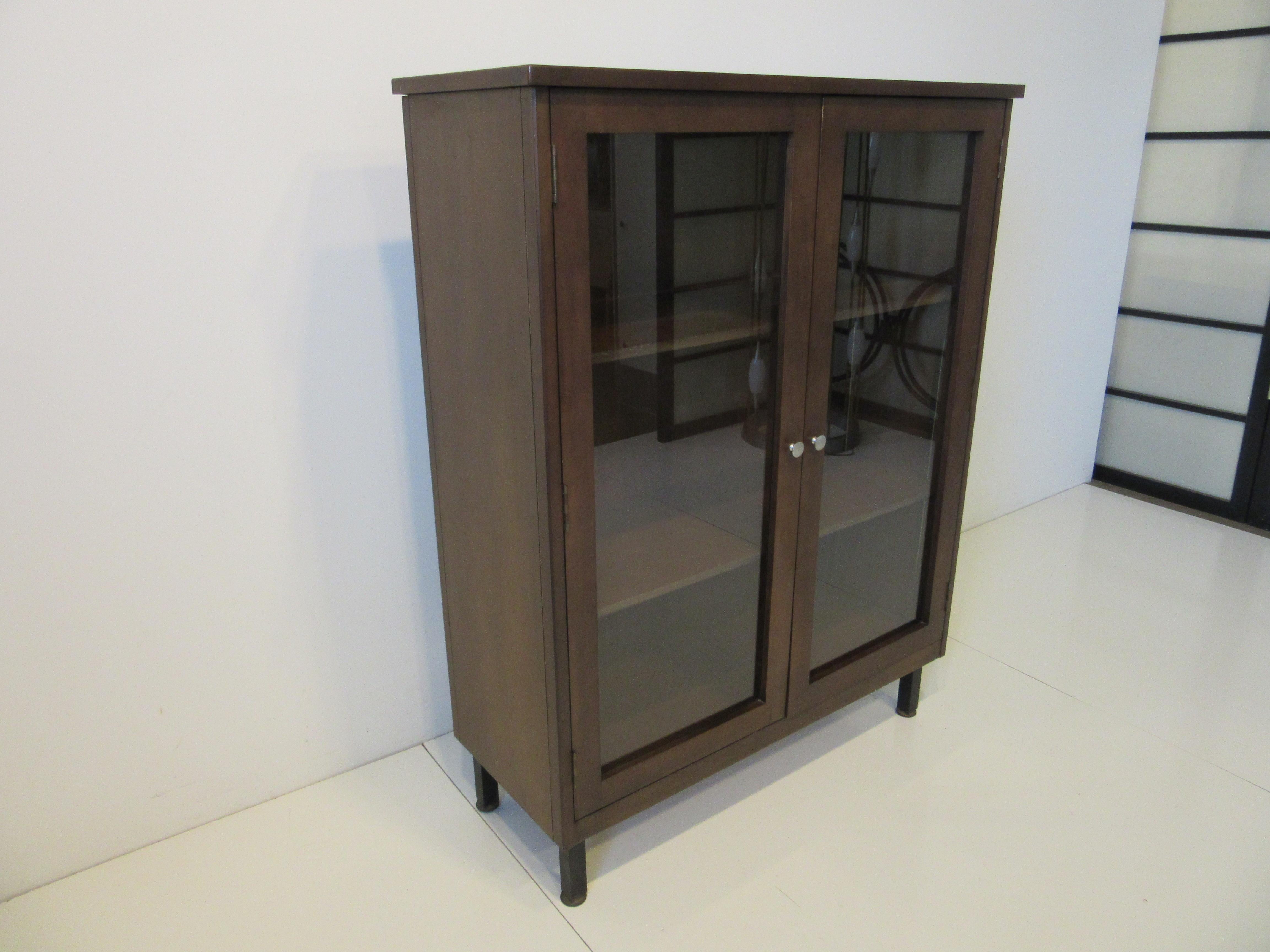 A glass framed two door walnut bookcase with one fixed shelve and one adjustable shelve with doors having brushed aluminum button pulls. This sturdy bookcase sits on satin black square tubed steel legs with adjustable foot pads, the top of the case