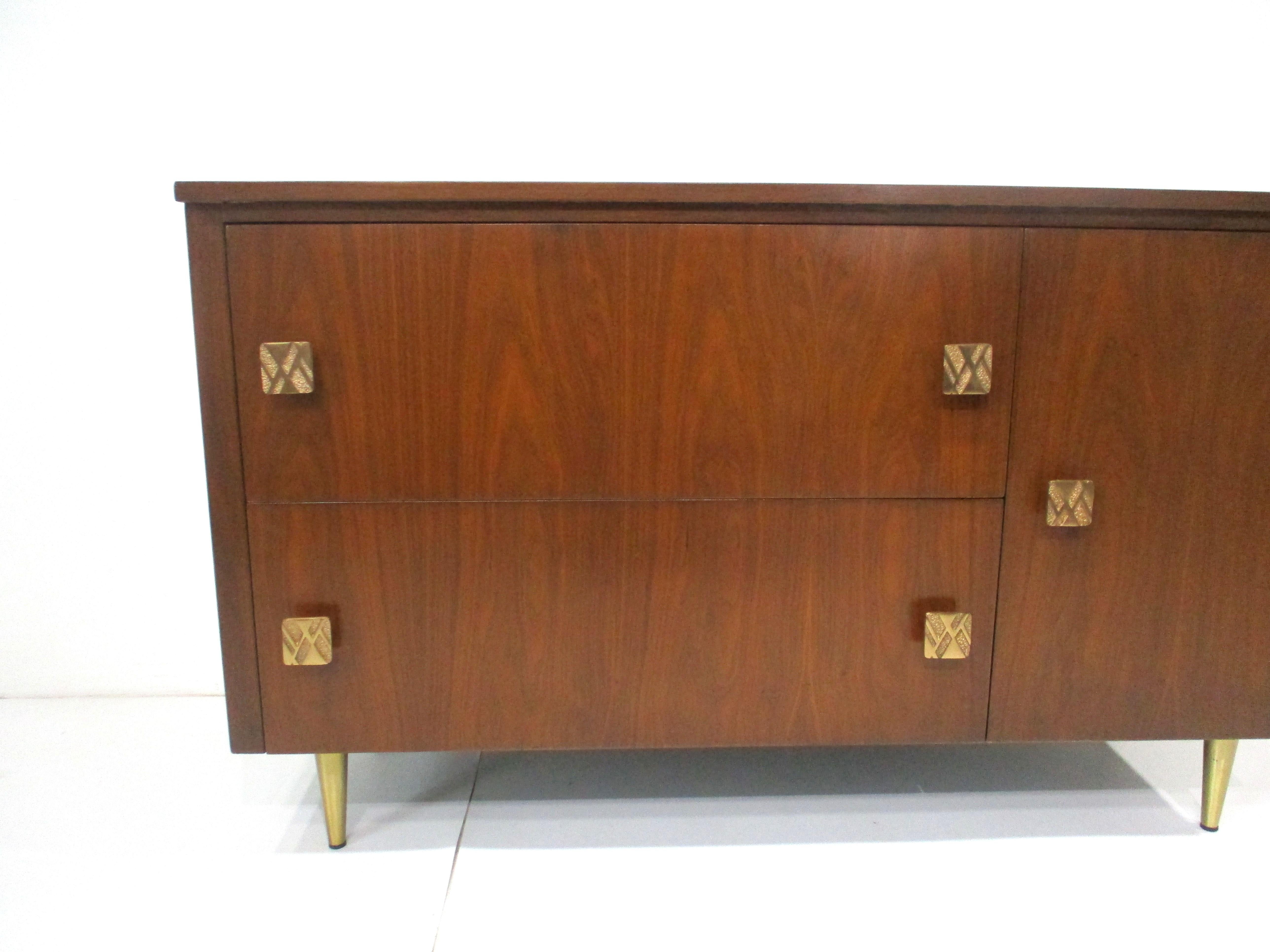 A medium dark walnut cabinet with wonderful graining , cast brass pulls and tapered brass legs . With two large deep drawers and a door with a non adjustable small half shelve the sculptural pulls having angular designs give the piece fine detail