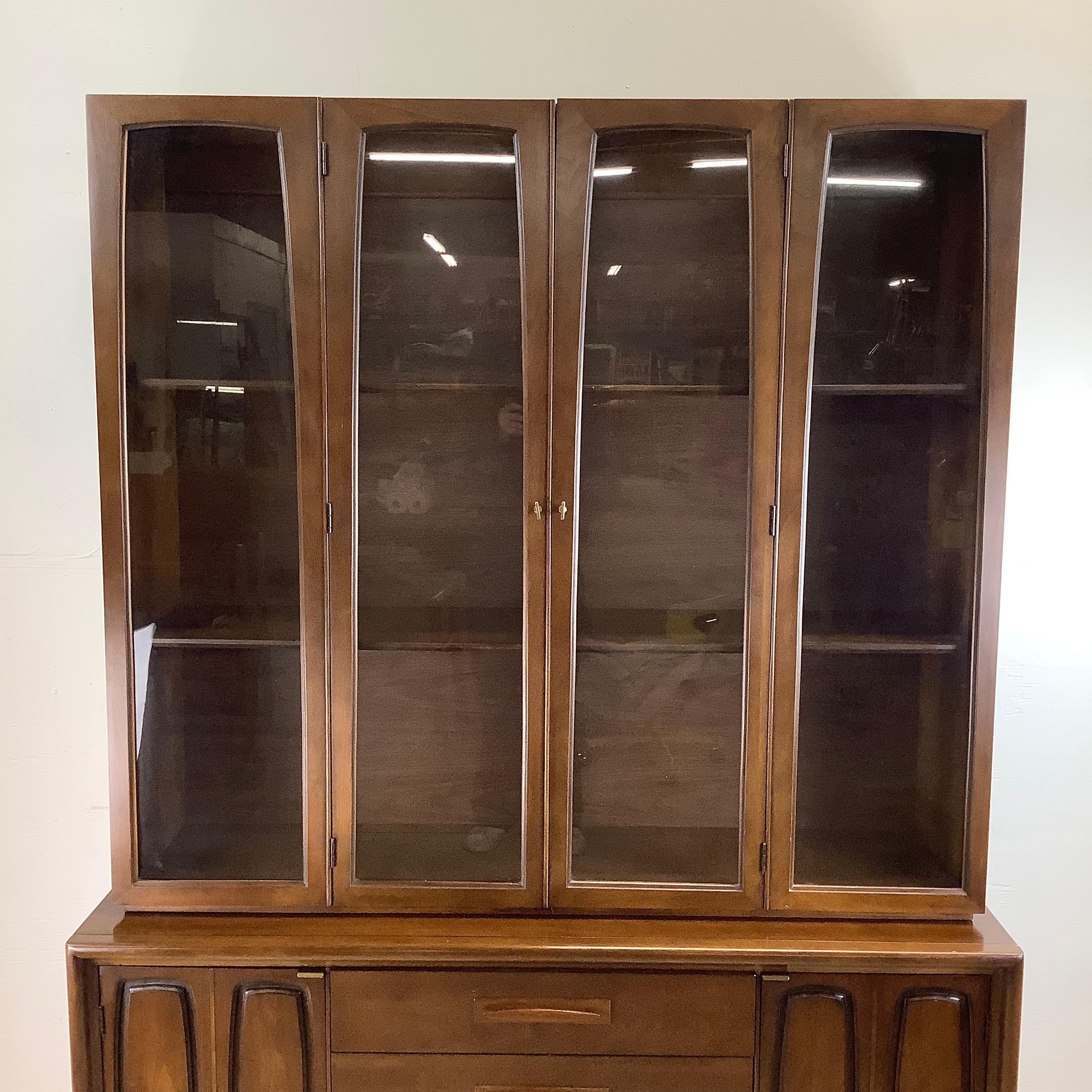 Add a touch of mid-century modern elegance to your dining space with this stunning Broyhill Emphasis Two-Piece Sideboard and China Cabinet. A masterpiece of both form and function, this versatile unit pairs a sleek lower credenza with a