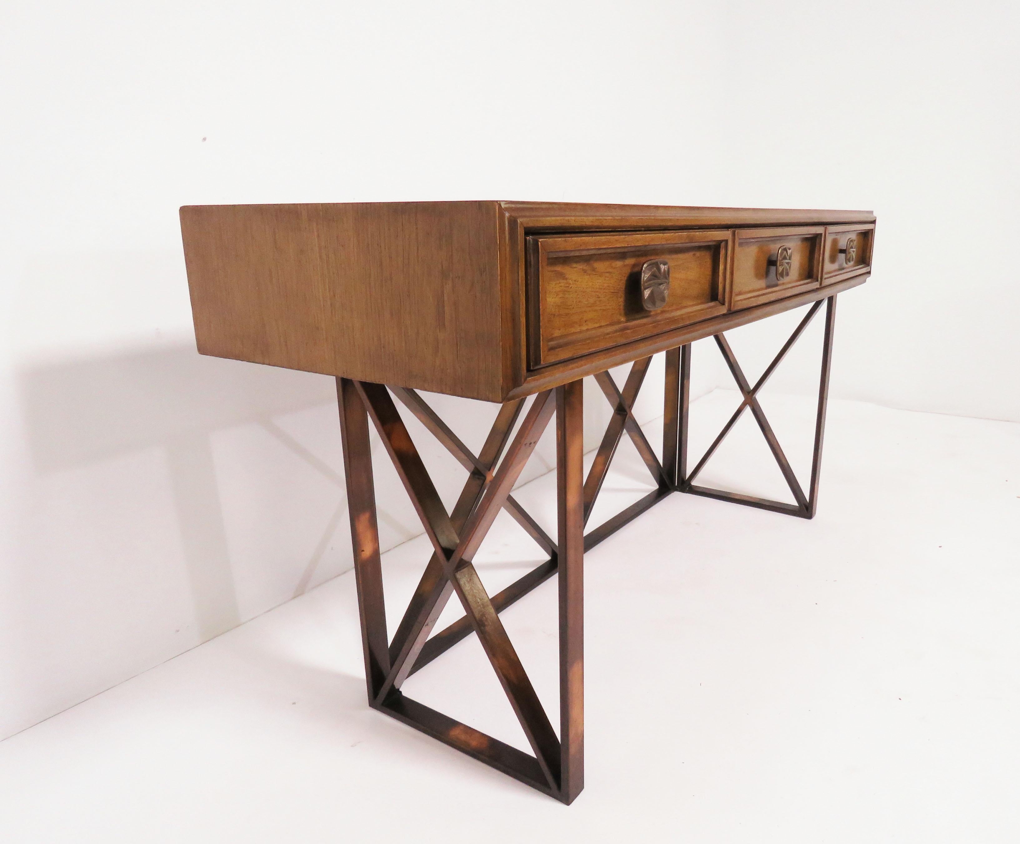 An American midcentury walnut console table of three drawers with modernist bronzed pulls, supported on an X-form caged bronzed base with copper highlights. At desk height (29”), this would work well as a hallway or foyer table. Measures: 56” long,