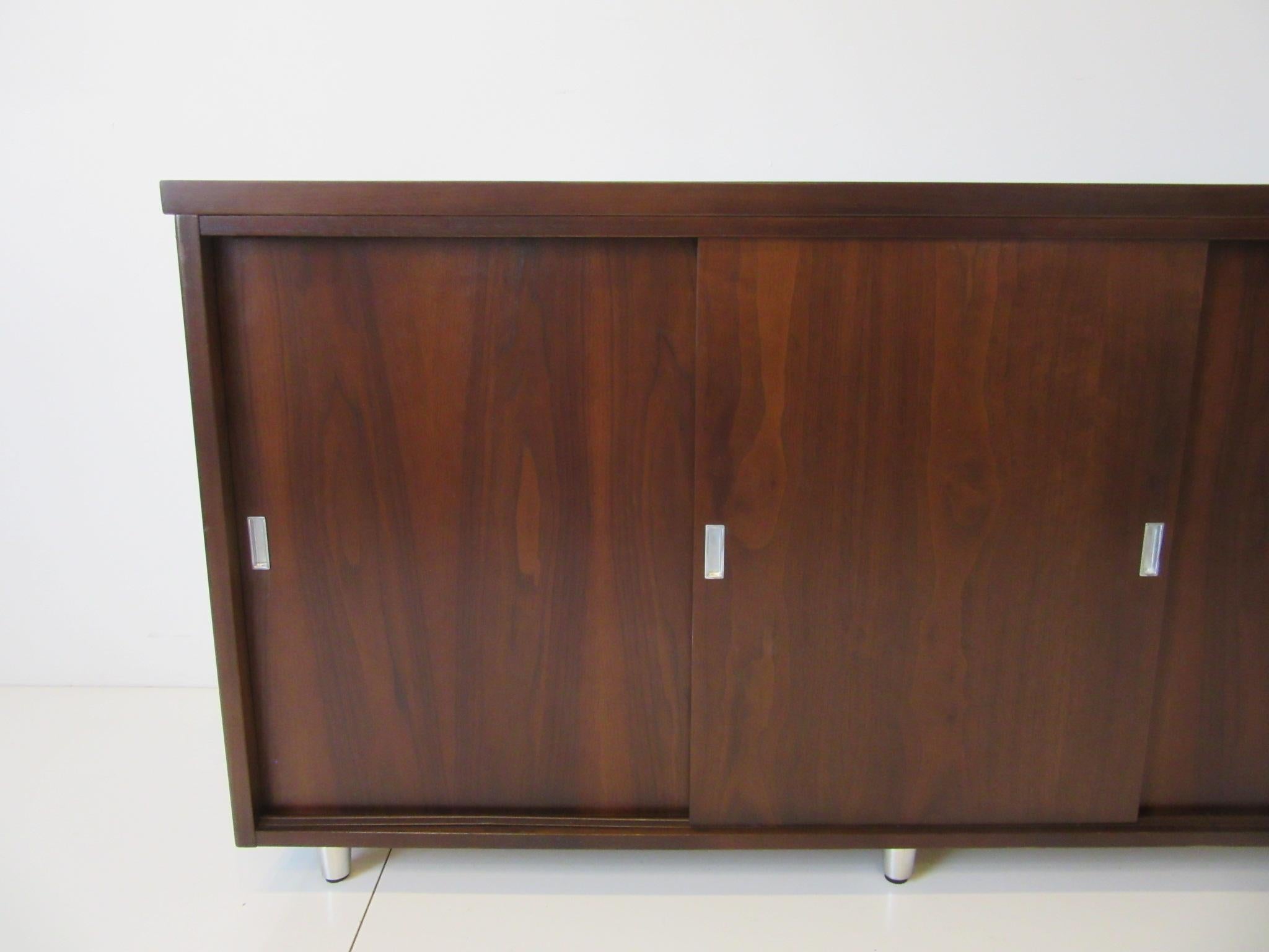 A dark walnut three sliding door credenza with four storage compartments, aluminum pulls and brushed aluminum legs having adjustable foot pads. The piece has a higher profile which is hard to find in a midcentury credenza, very well constructed
