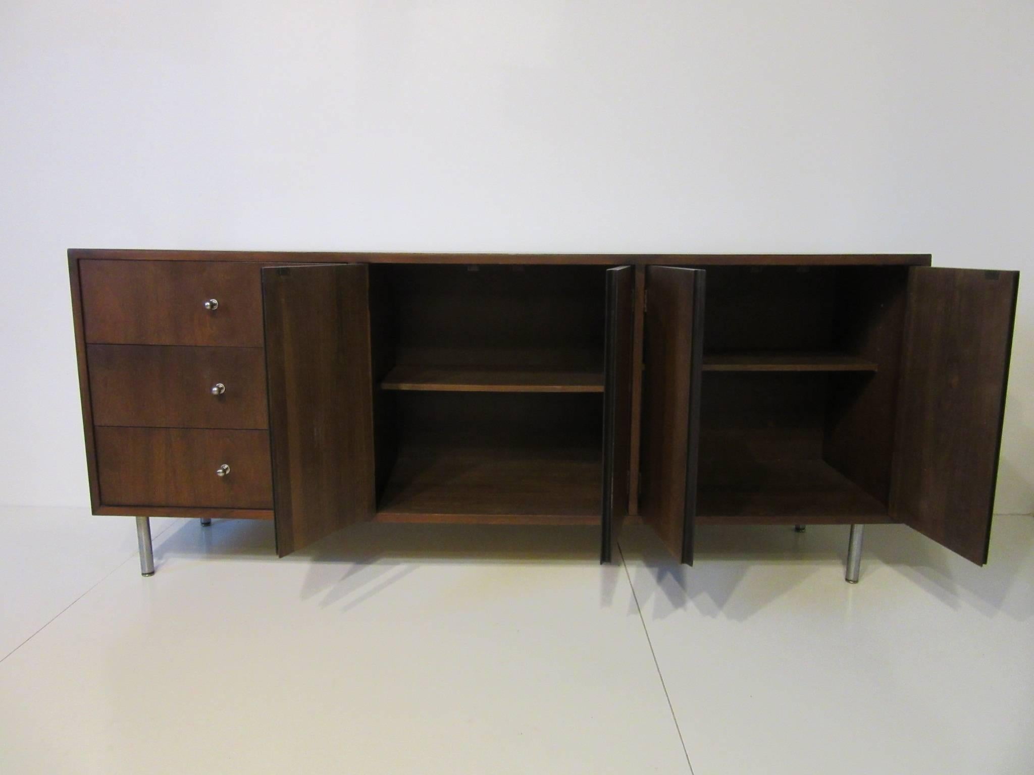 A dark walnut midcentury credenza with four doors, two adjustable shelves and three drawers with brushed stainless pulls that match the legs in the manner of George Nelson.