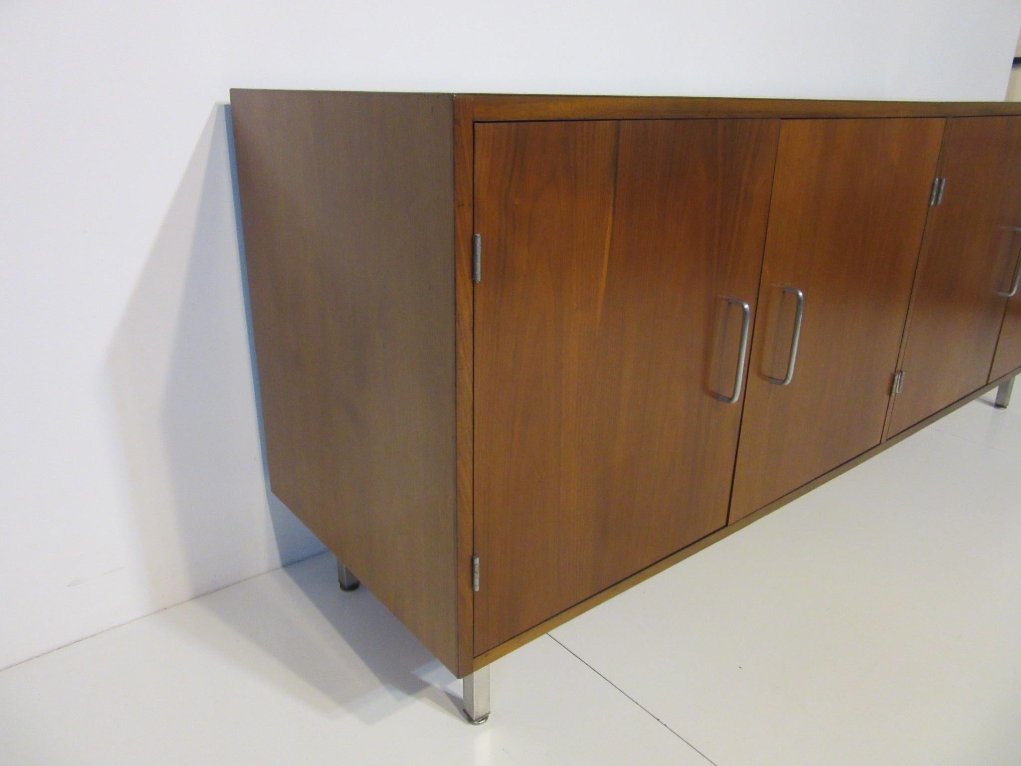 A larger four-door walnut credenza with stainless handles and legs and adjustable shelves, the backside has a satin black finish. In the manner of the Jens Risom furniture company.