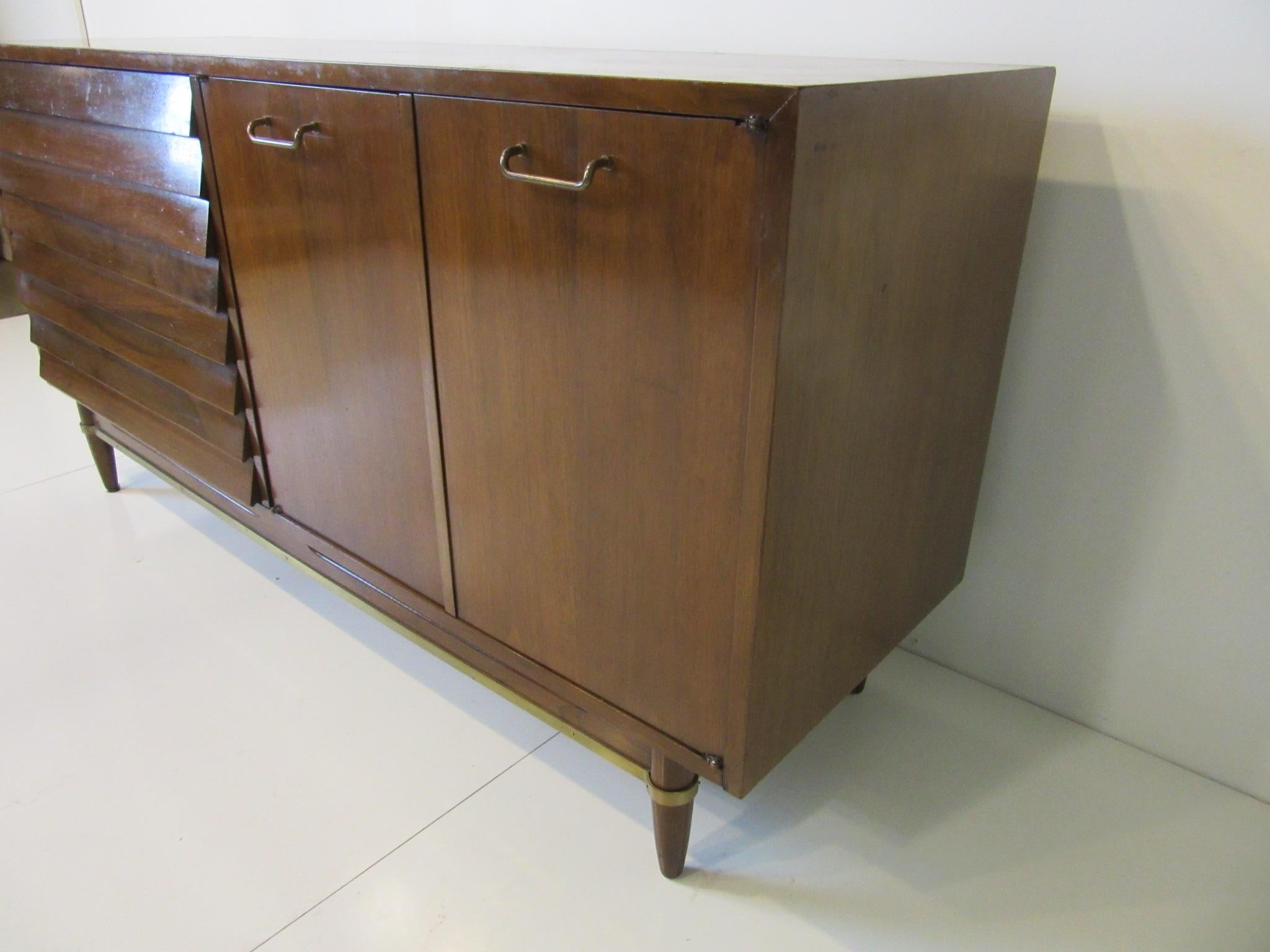 A dark walnut credenza / sideboard with double doors and three shelves, two cubby storage areas, and the other side having three large drawers with louvered fronts. Brass handles and lower matching stretchers with conical legs complete the