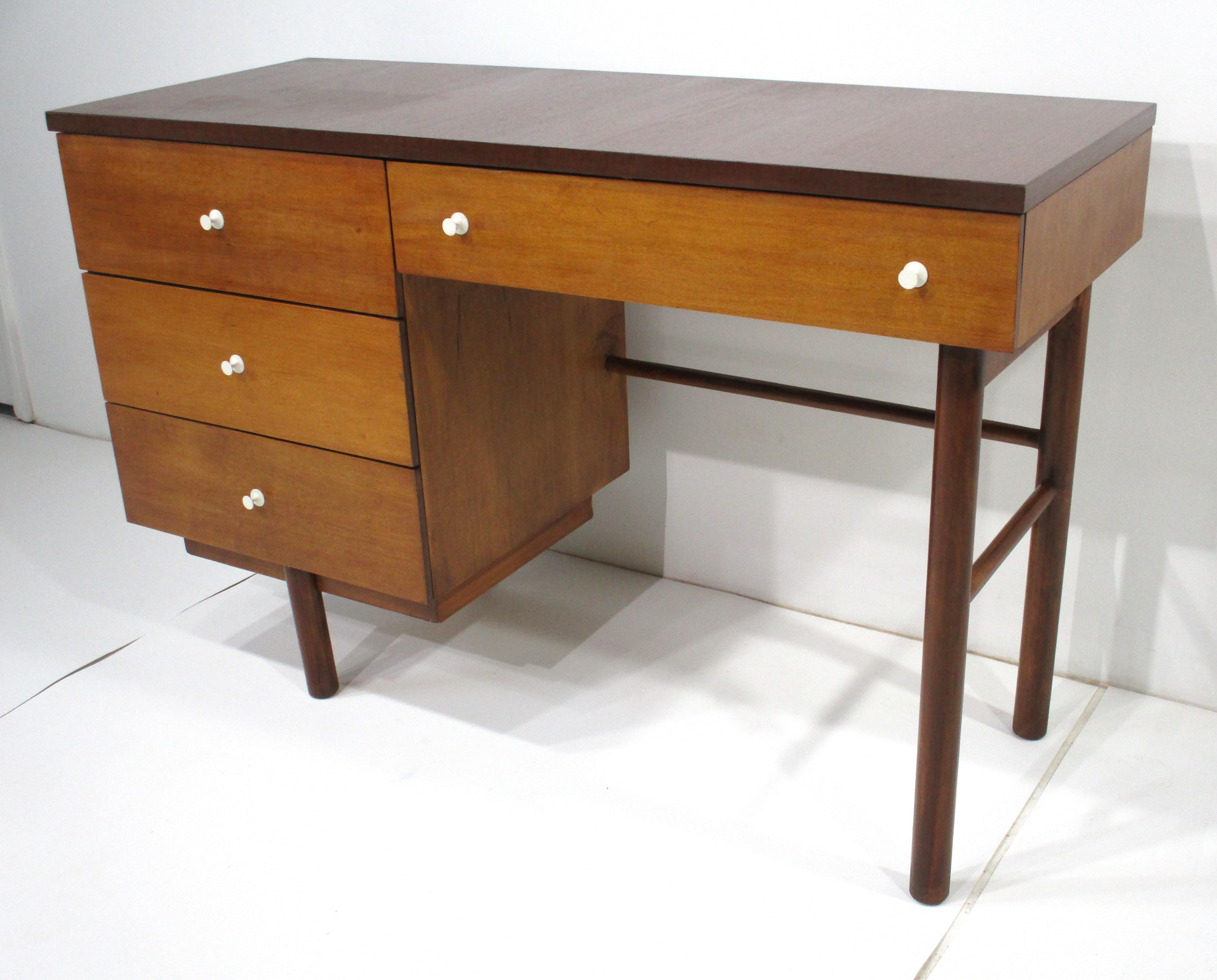 A writing desk with four light walnut drawers having tapered white pulls and a darker walnut frame and top . The simple aesthetics and combination of tones lets the piece work in many different interiors and mix with many styles , manufactured by