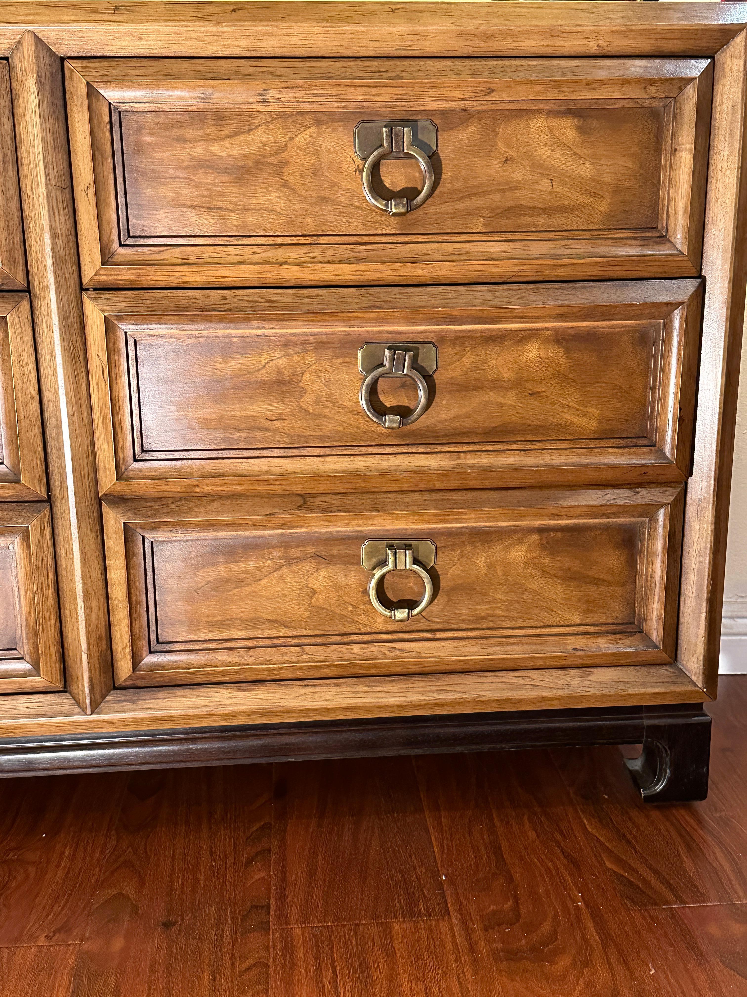 This Thomasville nine-drawer dresser features a medium wood finish with original brass drop-ring hardware. It rests on a black base with Ming-style feet, reflecting an Asian-inspired design. The dresser is in great condition and offers generous