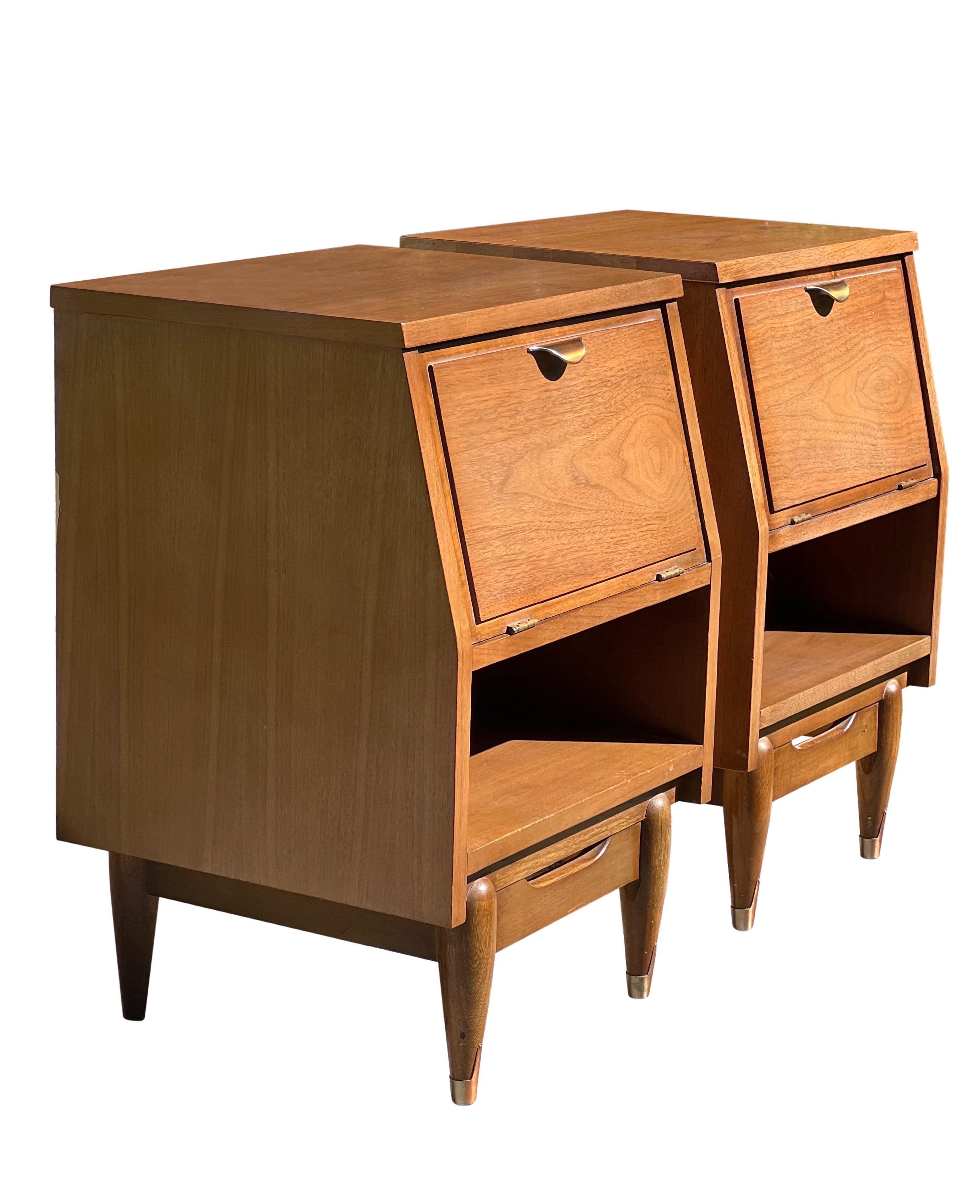 Handsome pair of Mid Century walnut nightstands.

The nightstands feature a drop-front cabinet door with a brass pull and open storage beneath. Tapered legs and decorative brass sabots. A super sturdy pair with great style and a unique silhouette.