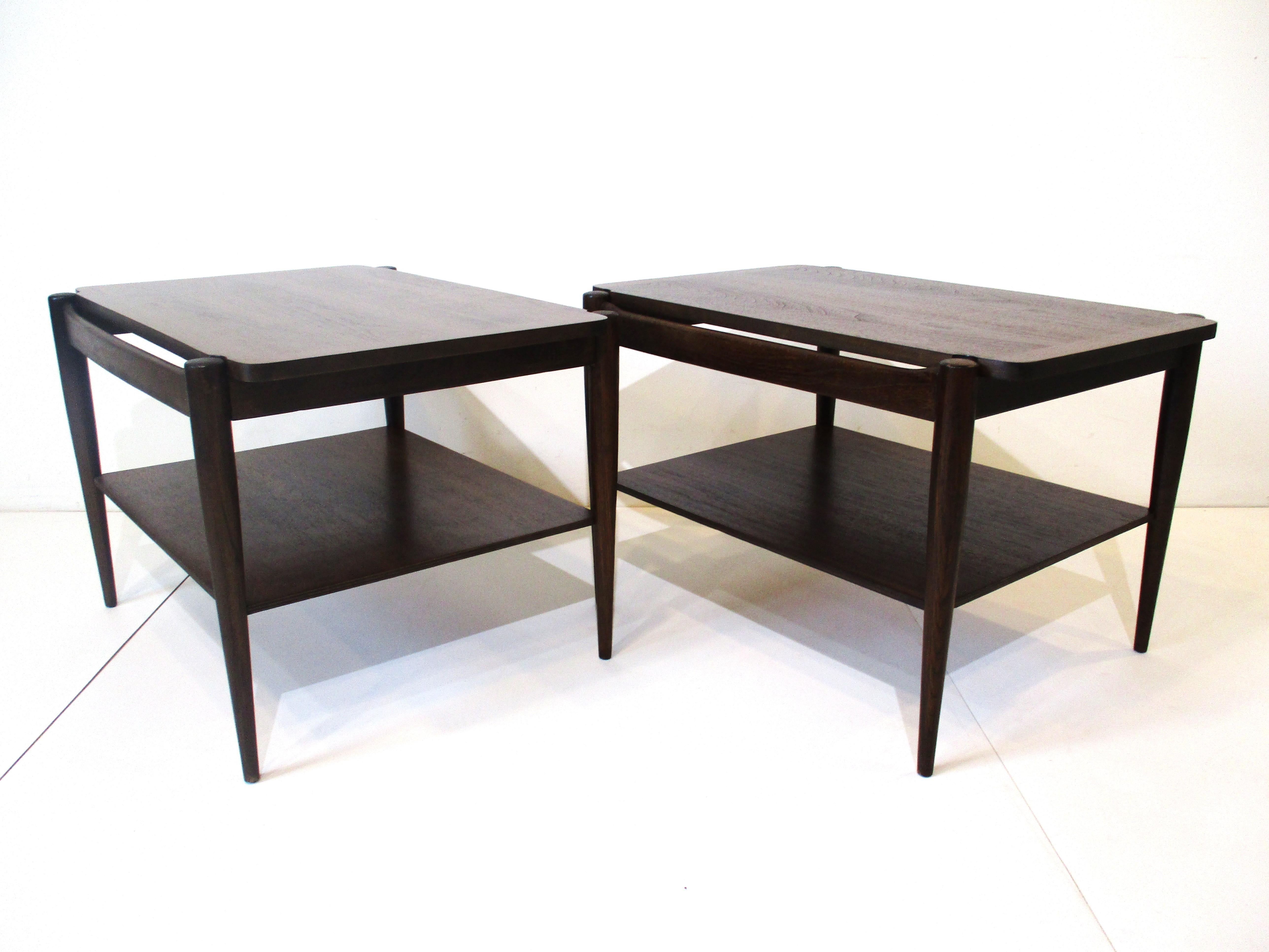 A pair of dark walnut end tables with lower shelve and long thin legs making for a light feeling . The top has wonderful graining with cut out corners for the legs in the style of Grete Jalk and danish modern . Manufactured by the Bassett furniture