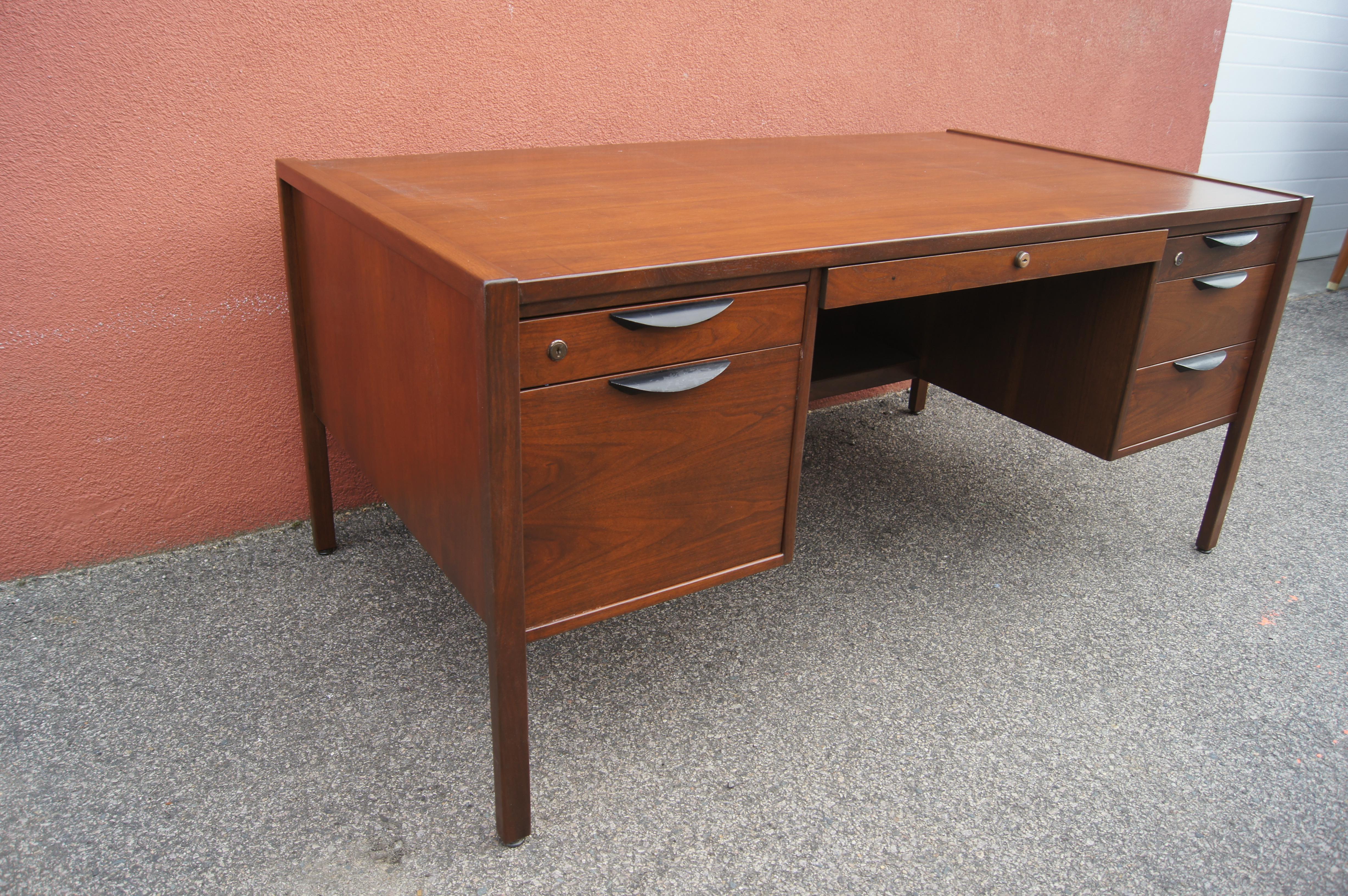 This executive desk, designed by Jens Risom in the 1960s, comprises a handsome walnut case that has been finished on all sides. Deep drawers with metal demilune pulls — two on the left, three on the right — offer substantial storage. A shallow