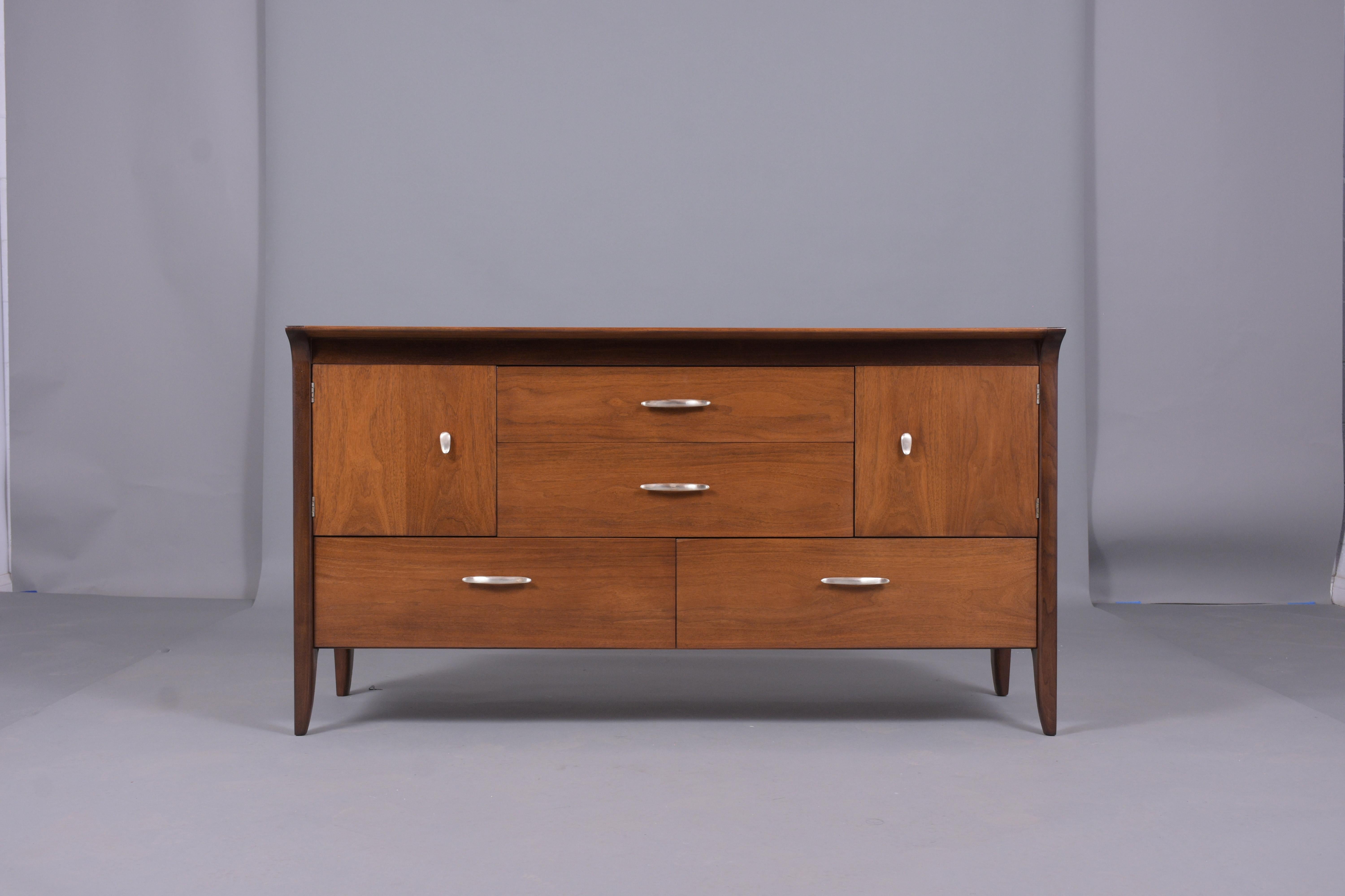 This vintage mid-century chest of drawers is crafted out of walnut and has been professionally restored by our team of in-house craftsmen. The dresser has been newly stained/lacquered finish, features four pull-out drawers, two doors with cubby