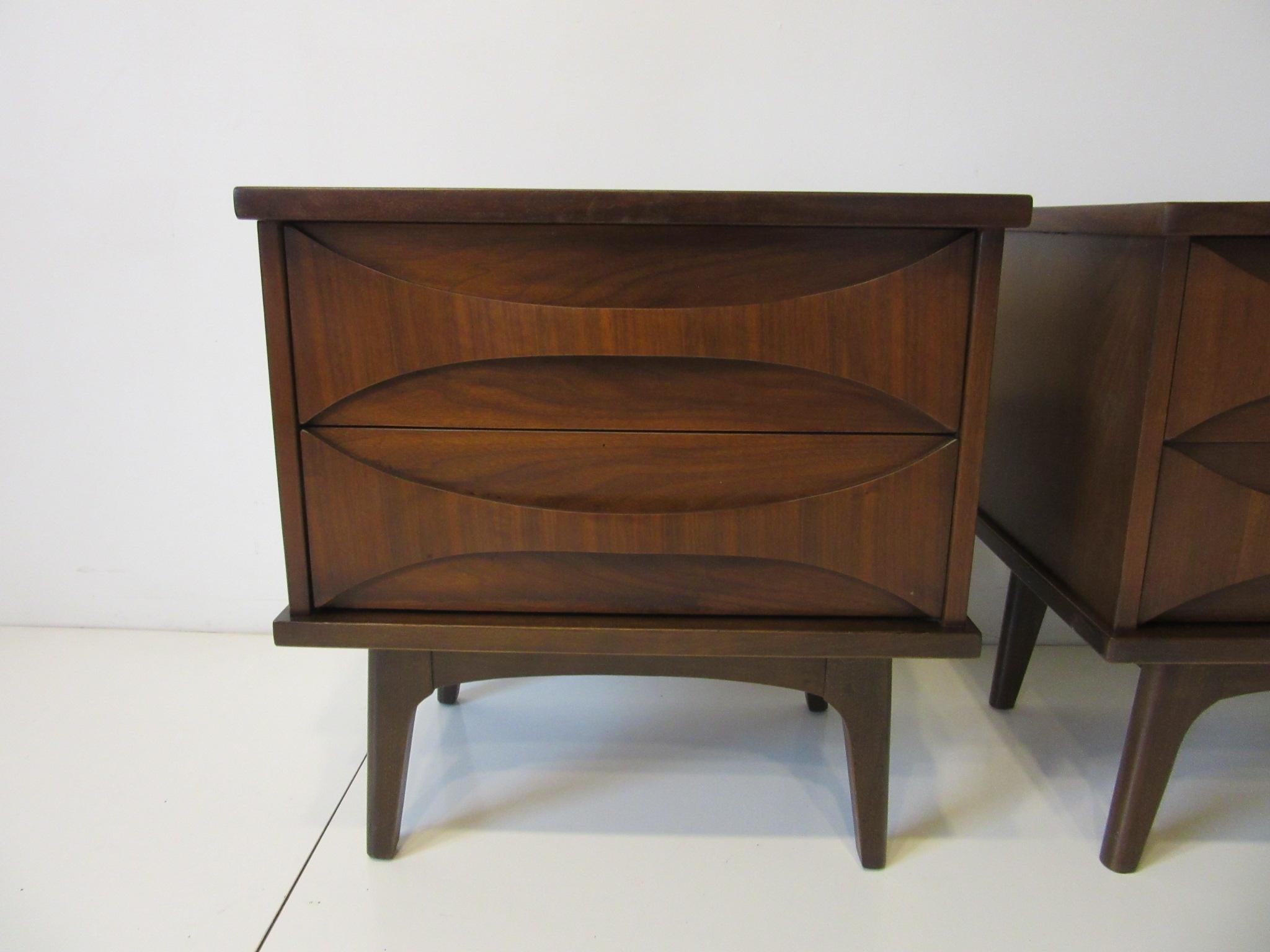 A pair of dark walnut nightstands with two drawers having cat eye styled fronts serving as pulls. Sitting on sturdy solid legs and styled in the manner of Brasilla and Perspecta furniture.
