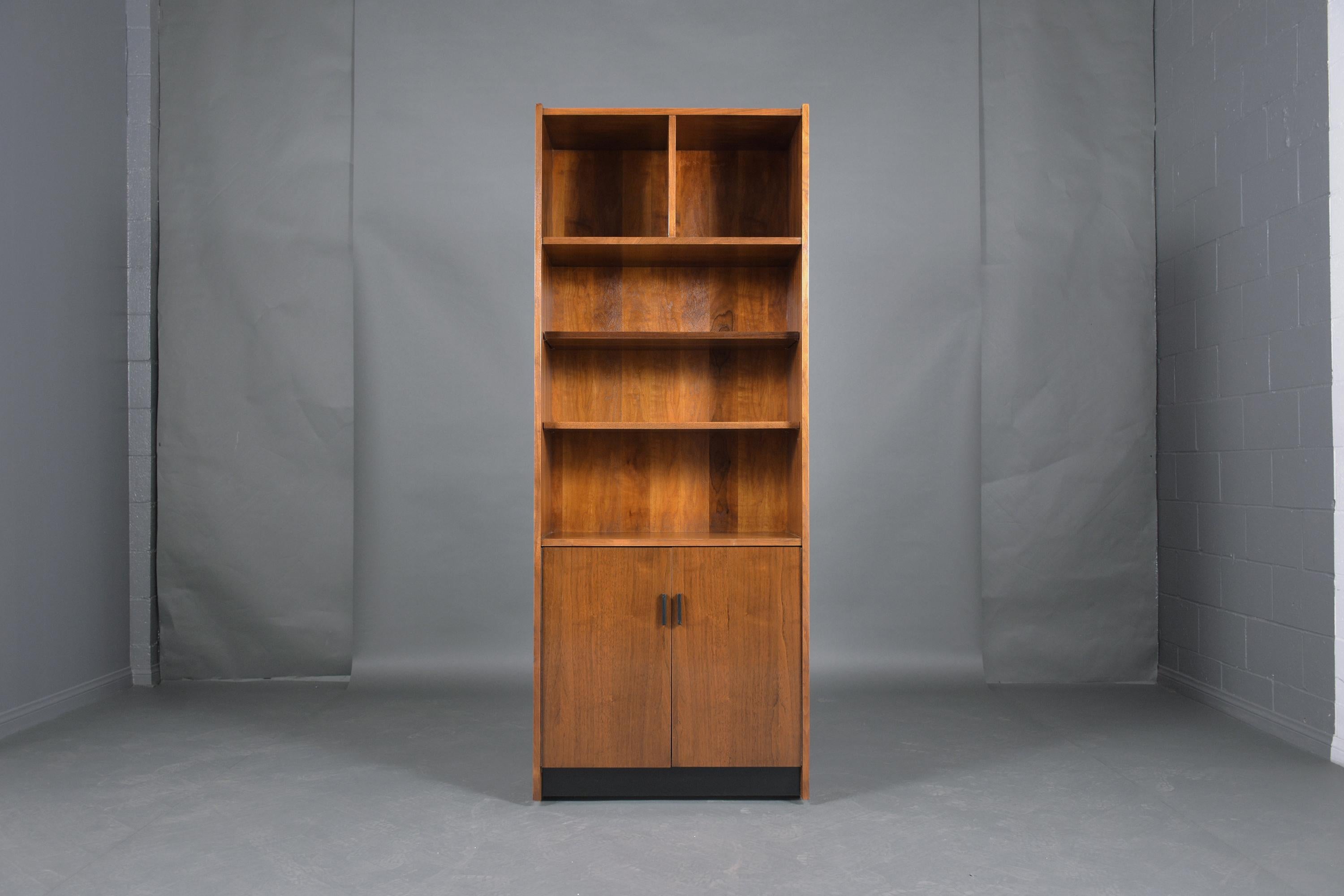 This vintage mid-century modern bookcase is crafted out of walnut wood and has been professionally restored by our team of in-house craftsmen. This wall unit features two cubbies and two open shelves on the top half and the bottom comes with two