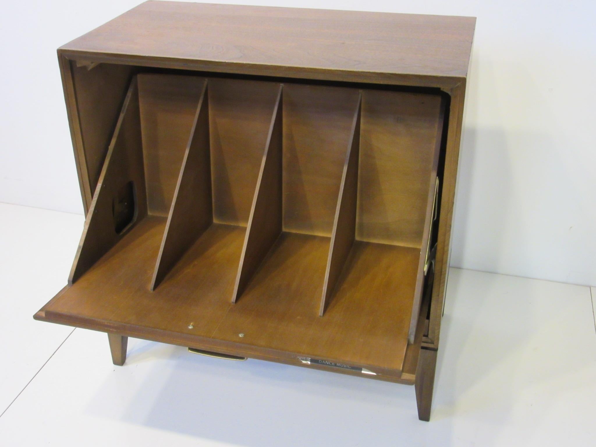 A dark walnut pull down front drawer midcentury record cabinet with four storage bins able to hold up to 130 plus albums. A brass handle to the detailed four walnut panel front and the weight of the records keeps the door firmly in place and hides