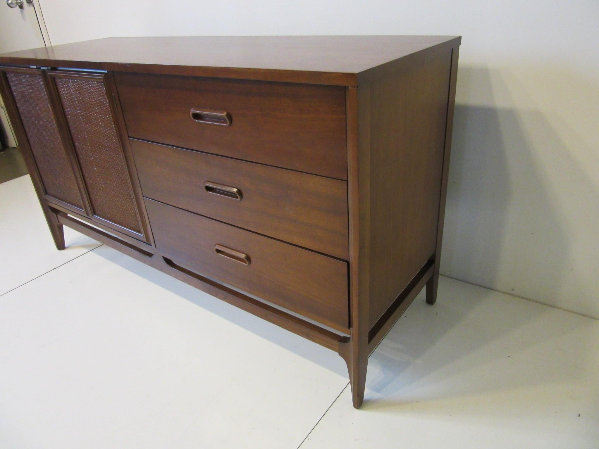 A smaller sized walnut credenza / server with three drawers having built in pulls and two doors and one shelve with storage. The double door fronts are woven with bronze toned upper pulls and wonderfully grained top.