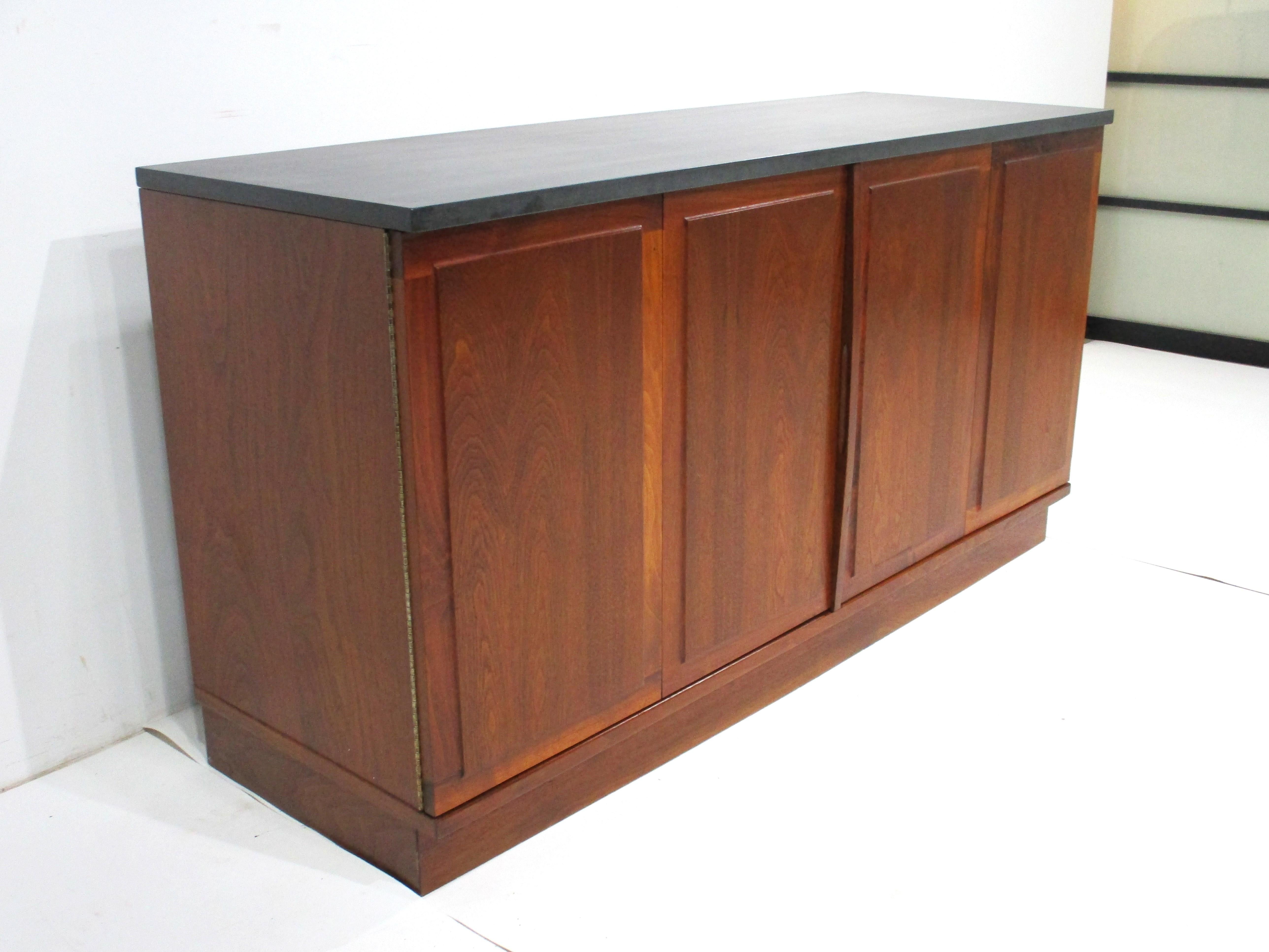 Una credenza di metà secolo in noce medio scuro, ben realizzata, con due porte a due ante con cerniere tipo pianoforte e due ripiani regolabili. Il piano in pietra di ardesia grigia conferisce al pezzo un tocco di stile ricco e pulito. Questa