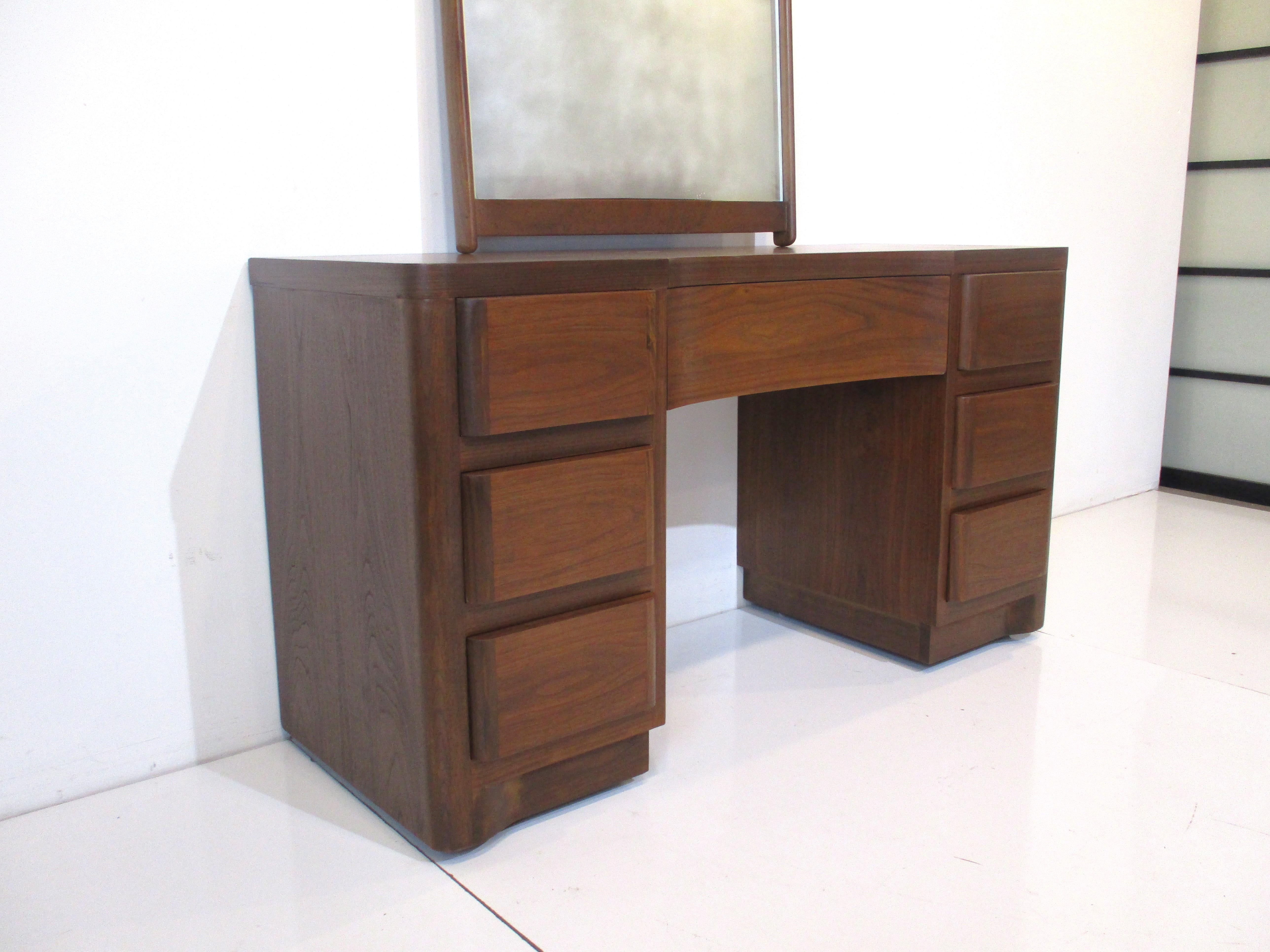 A midcentury walnut vanity with seven drawers one middle and three to each side well crafted in the manner of the Widdicomb furniture company. Having rounded drawer ends, wonderful graining to the wood and a nice cut out for your seating with a