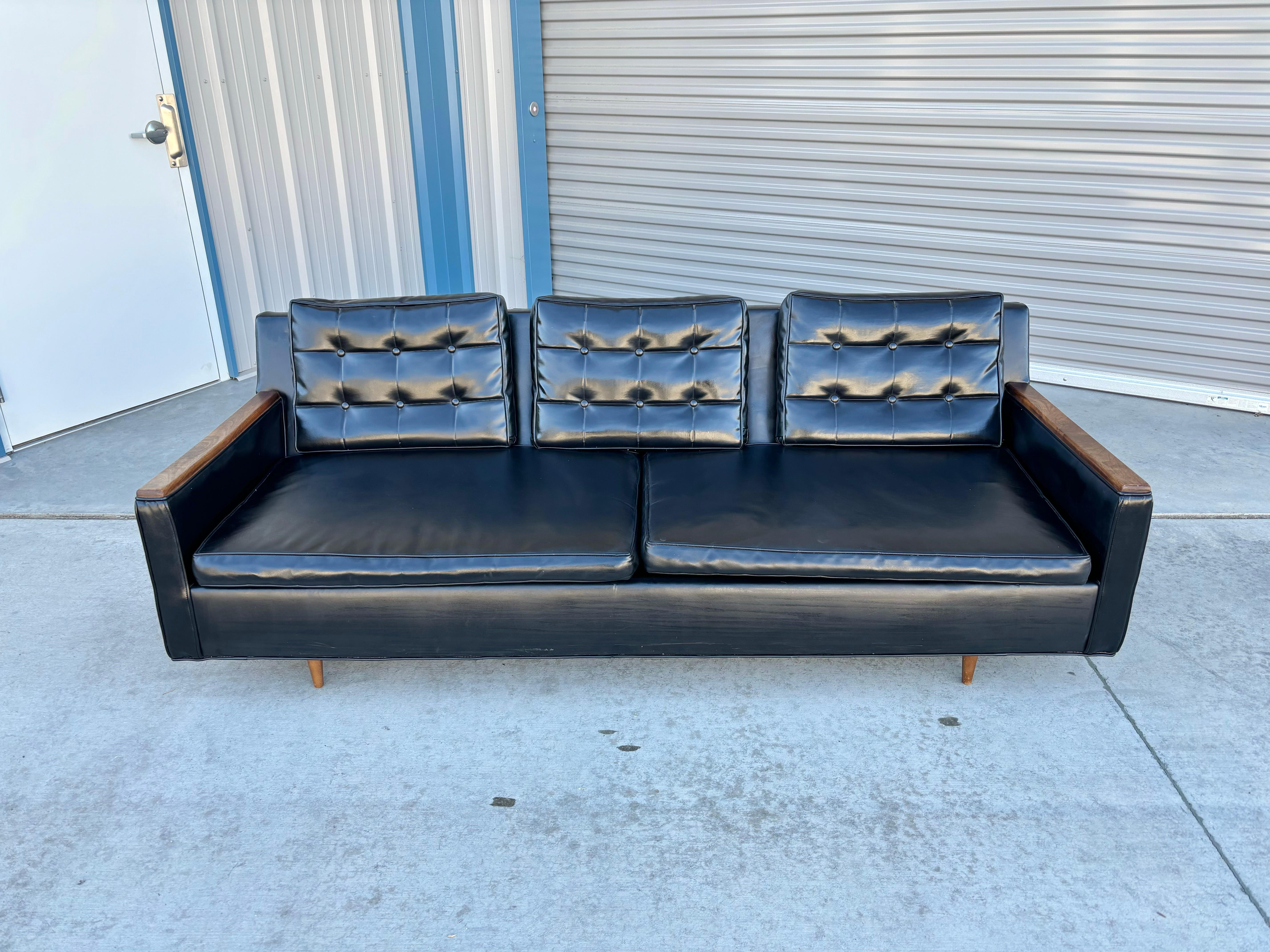This mid-century walnut and vinyl sofa was designed and manufactured in the United States in the 1970s. This magnificent piece of furniture boasts sleek black vinyl upholstery, and what sets it apart is the exquisite walnut slates adorning its