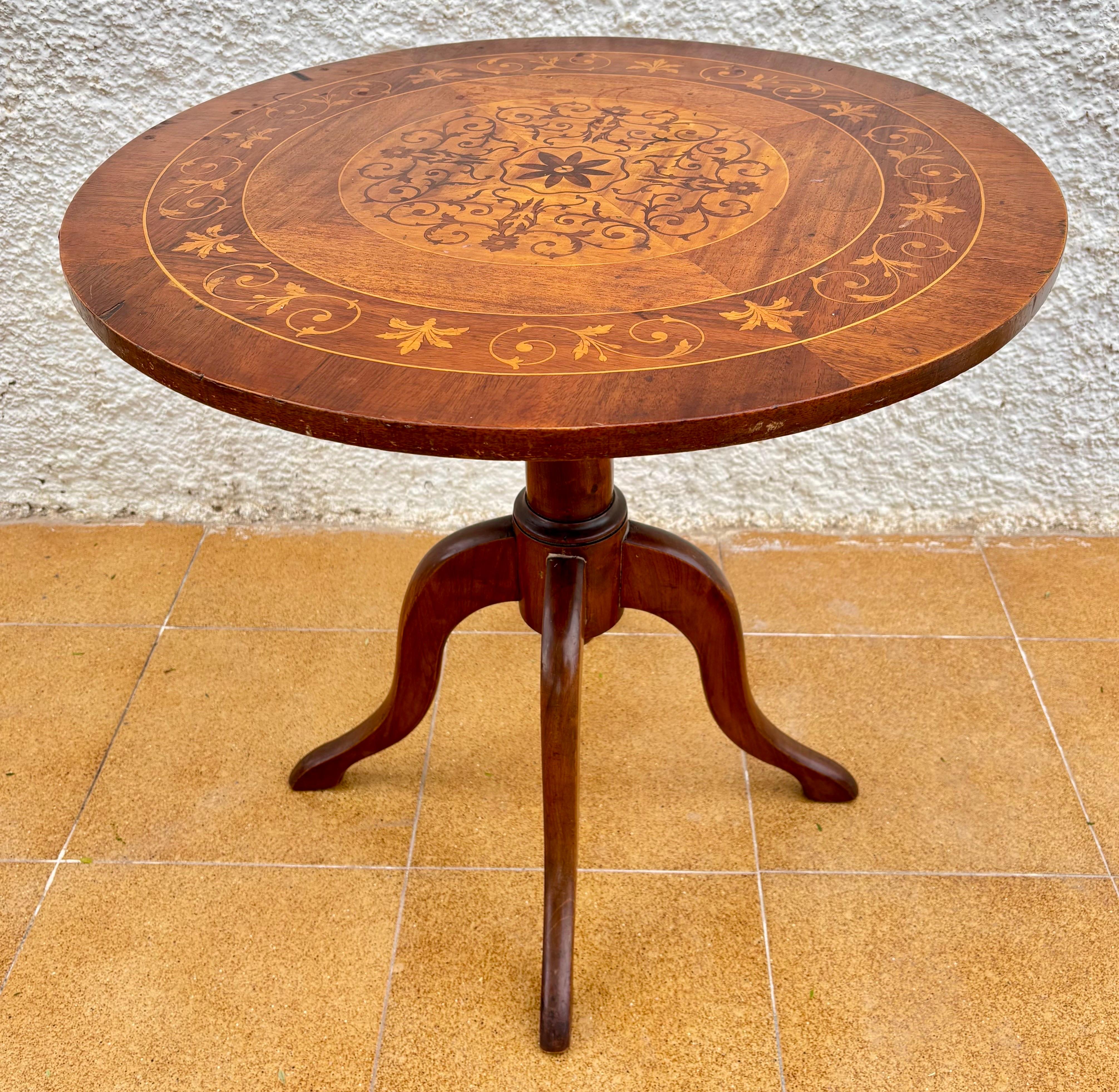 Magnifique table d'appoint ronde du début du 20e siècle, avec un plateau en marqueterie ornée d'incrustations florales et géométriques. Le plateau circulaire présente une combinaison raffinée de bois clairs et foncés, créant un médaillon central