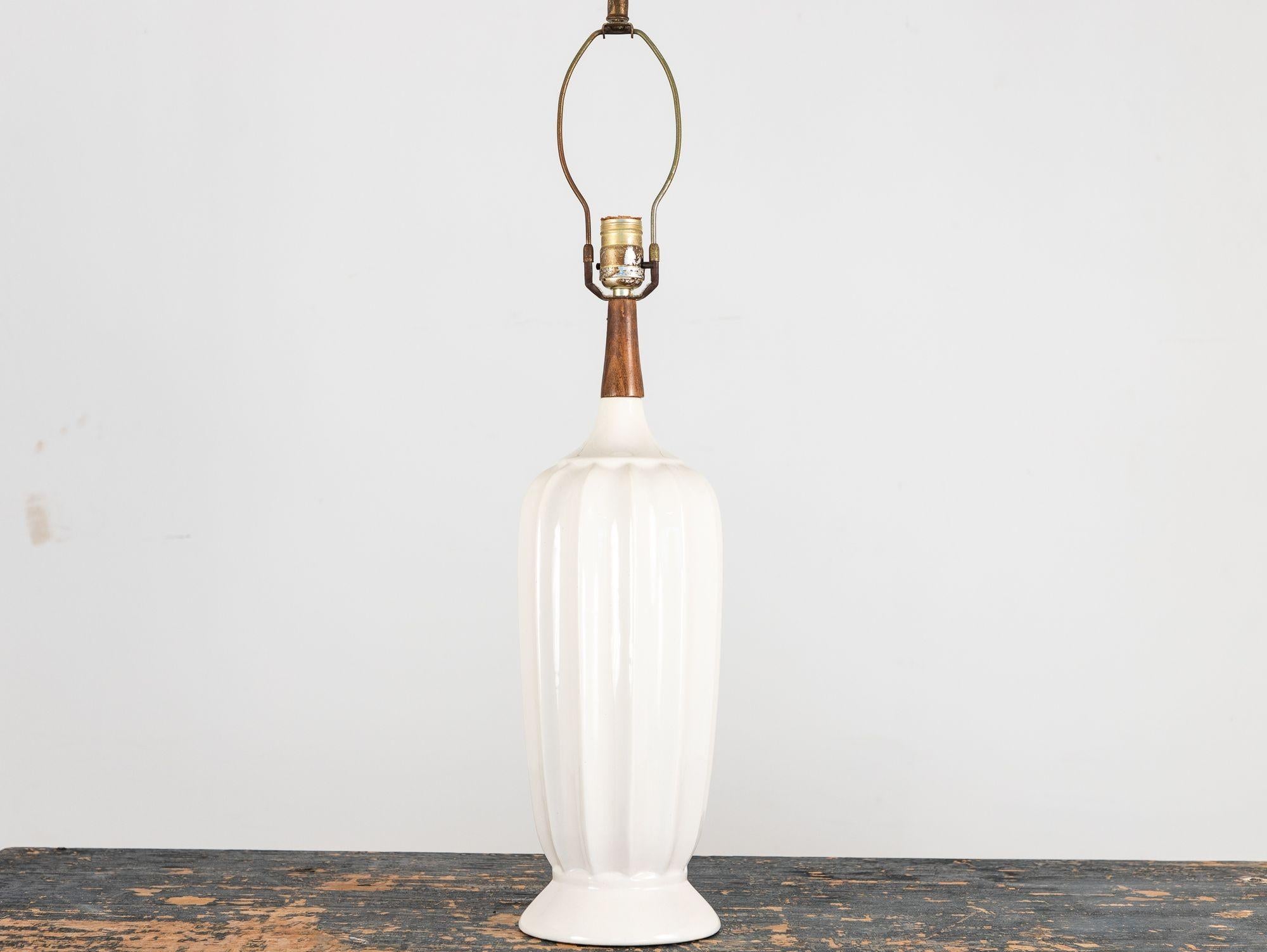 Cette élégante lampe de table vintage des années 1960 présente un corps cannelé en céramique blanche avec une forme légèrement effilée, s'élargissant vers le haut et s'évasant à la base pour une silhouette intemporelle. Le long col en bois ajoute de