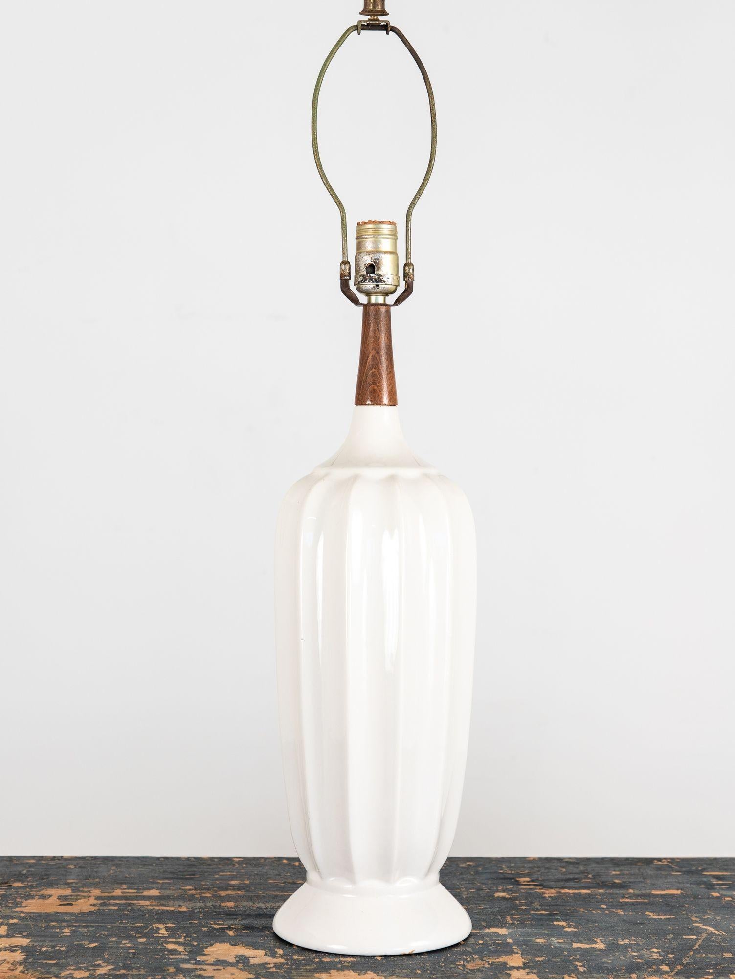 Cette élégante lampe de table vintage des années 1960 présente un corps cannelé en céramique blanche avec une forme légèrement effilée, s'élargissant vers le haut et s'évasant à la base pour une silhouette intemporelle. Le long col en bois ajoute de