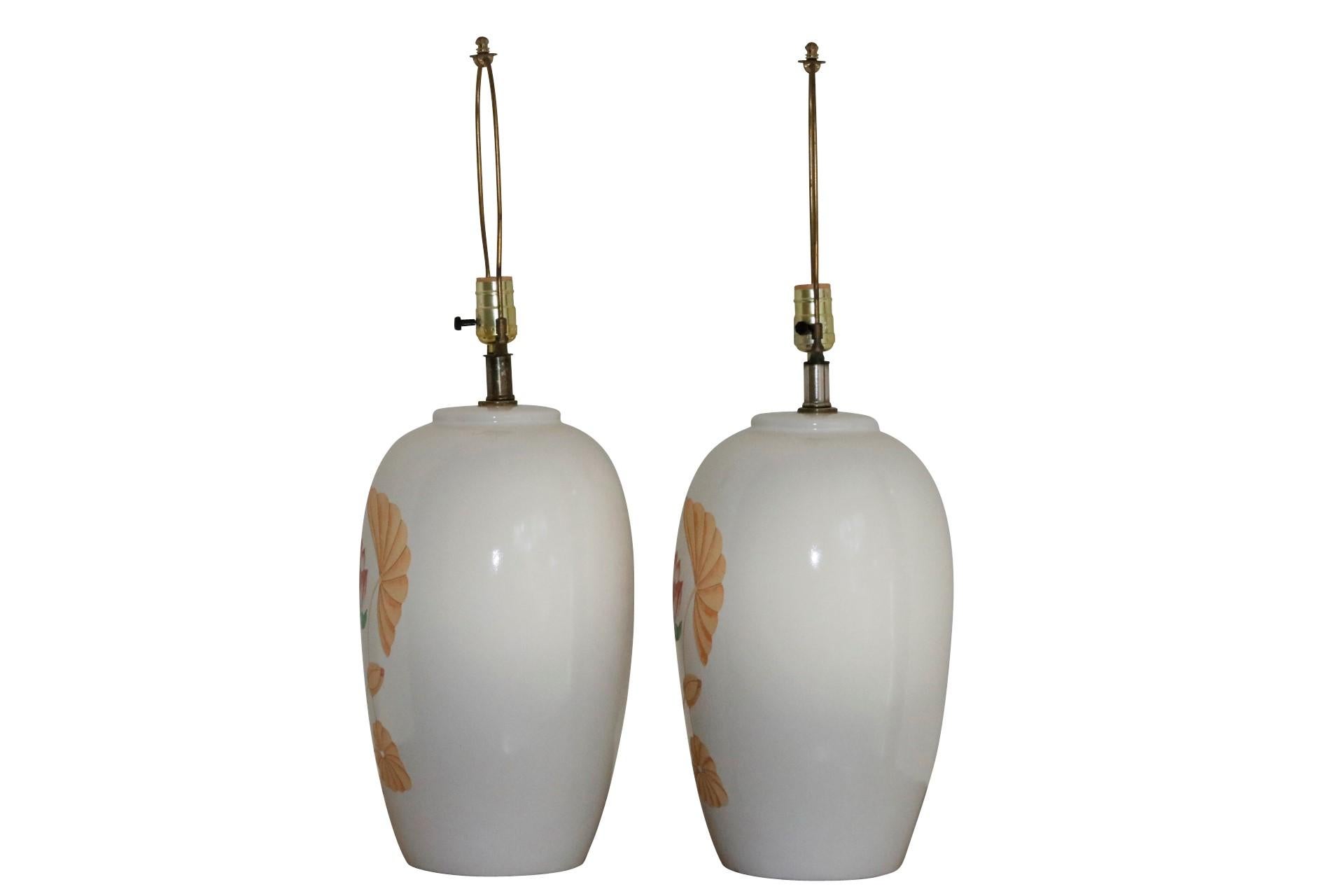 A pair of white ceramic table lamps. Fronts are decorated with a printed water lily motif in pink and green, framed with lilies in yellow. Wired and working. Each lamp measures 19.5