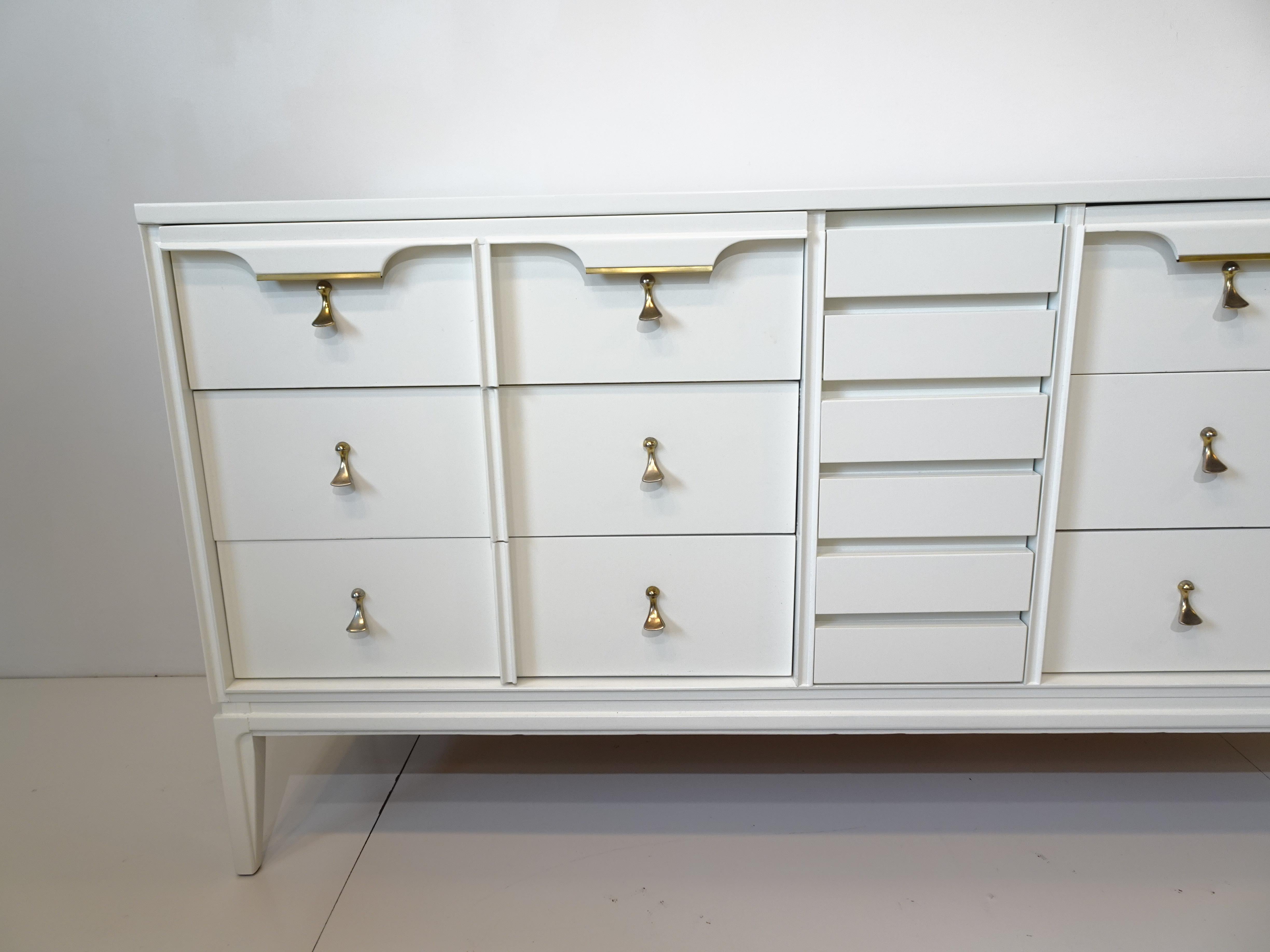 A satin white mid century dresser with six large drawers to each side and three smaller center drawers having brass trim and chrome pulls . A clean and simple form with scalloped styled top drawer trim retaining the manufactures branded mark by the