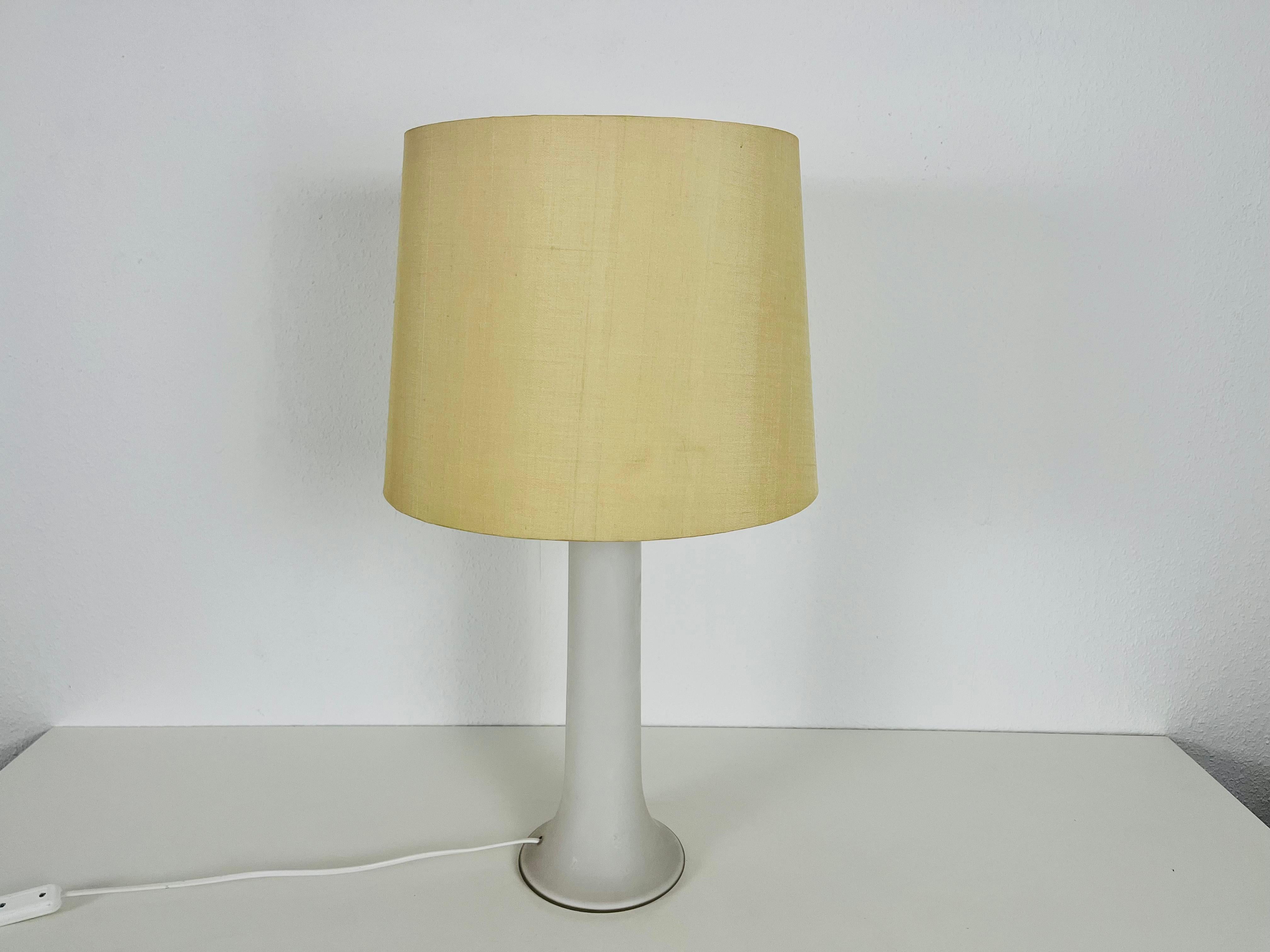 Eine schöne große Tischlampe aus den 1960er Jahren von Luxus Sweden. Der Sockel ist aus weißem Glas gefertigt. Der Lampenschirm ist aus Stoff gefertigt und hat eine beige Farbe.

Die Leuchte benötigt eine E27-Glühbirne. Funktioniert sowohl mit