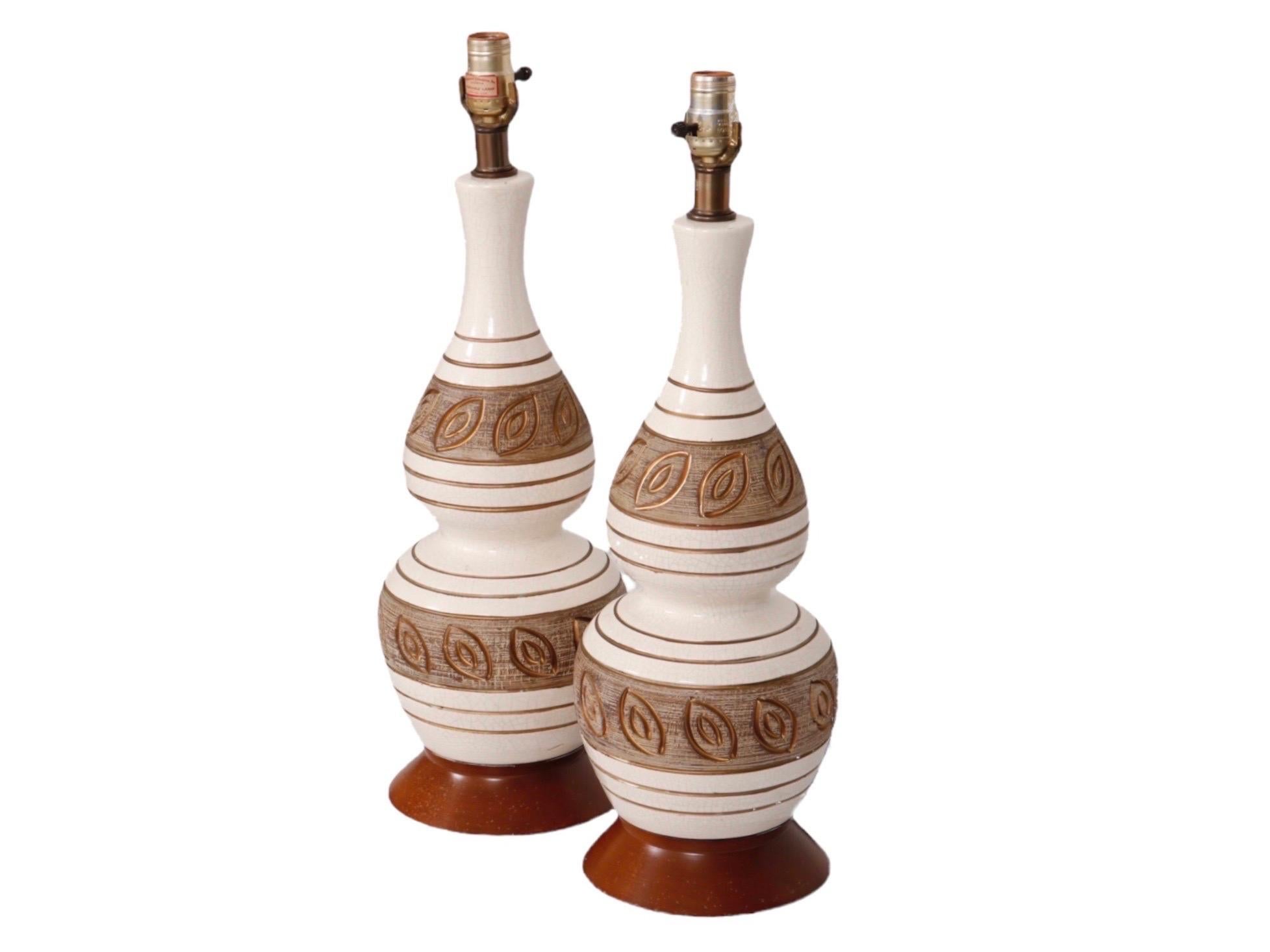 A pair of mid-century baluster shaped ceramic and teak table lamps. Finished in a white craquelure glaze, hand decorated with horizontal lines and a simple leaf motif in gold. The leaf pattern is set against a brown glazed background marked in such