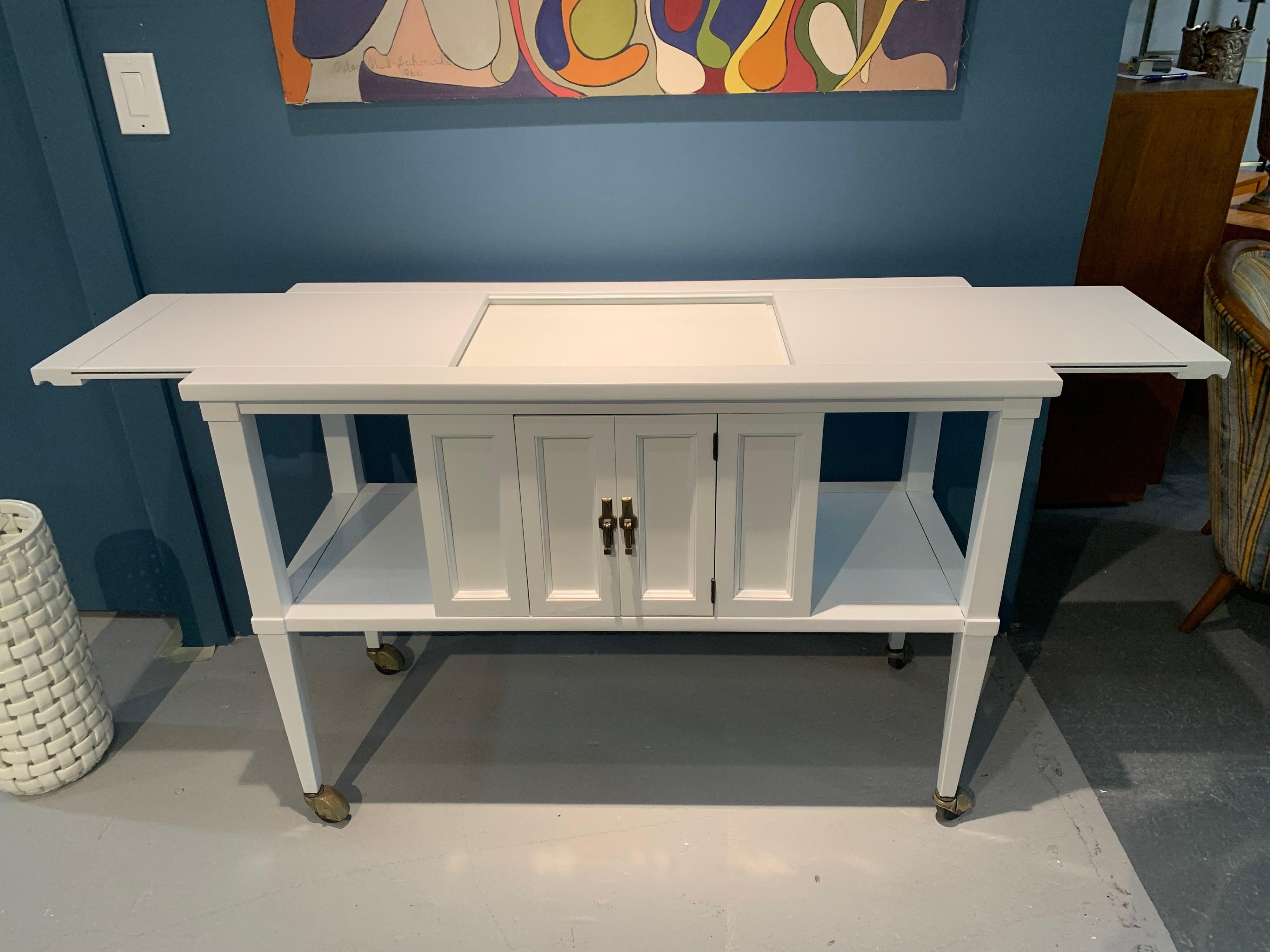 Mid century rolling bar cart buffet has been recently lacquered with a clean white finish. It has two tiers with center cabinet and pullout sides. Serve food or drinks in style with this bar cart that rolls to where you need it.