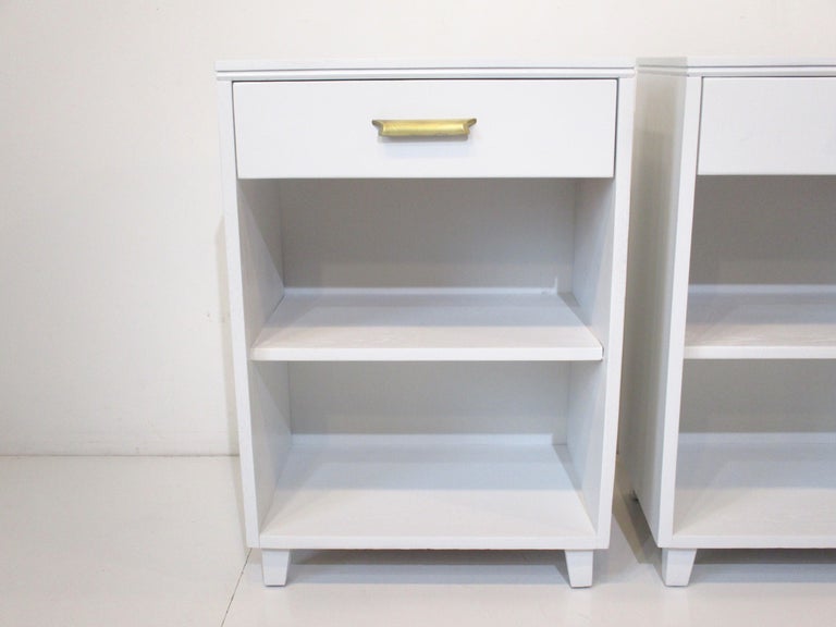 A pair of satin white mid-century nightstands in a taller profile working well with the new matrass heights . Having one upper drawer with a nice long brass pull and storage below with a non adjustable shelve, manufactured by the Oak Masters Modern