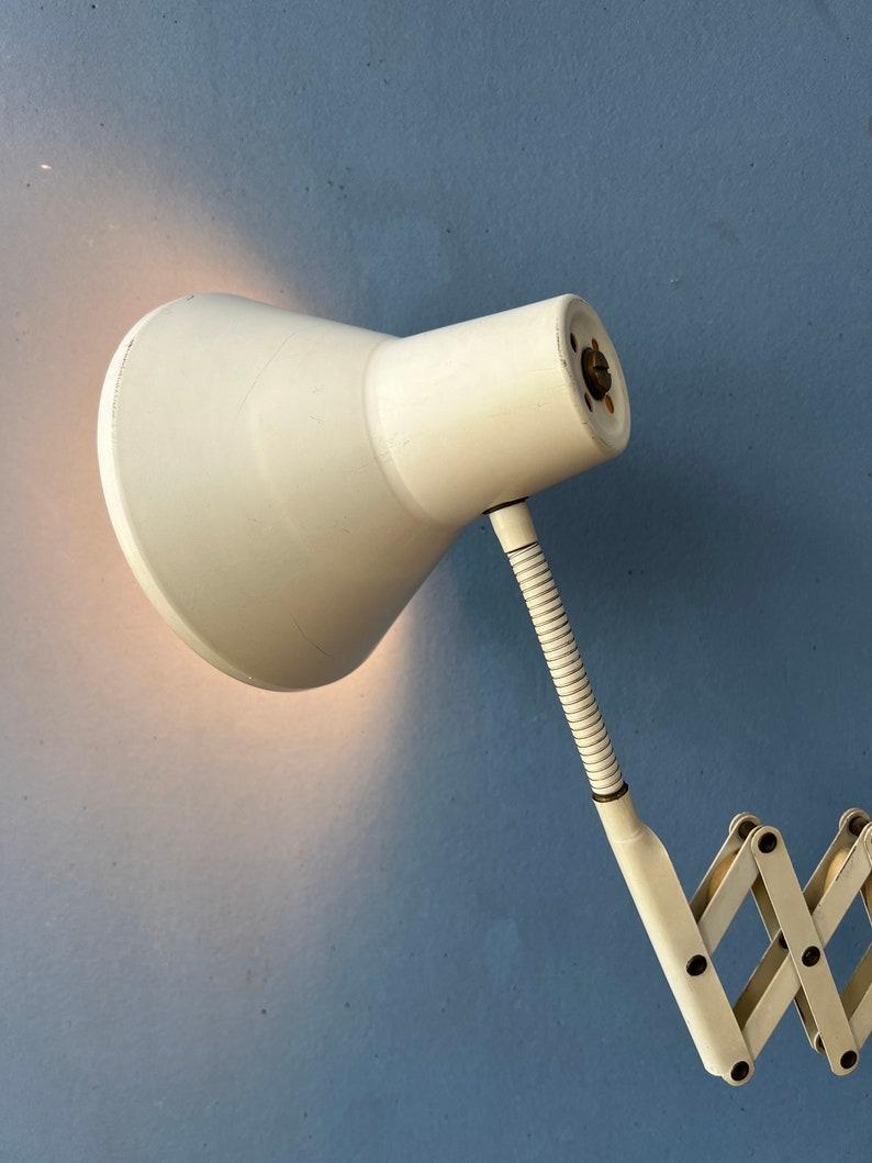 Mid century white scissor wall lamp made out of metal. The scissor arm allows you to adjust the lamp's position to suit your needs. Also the shade itself can be easily adjusted in position. The lamp requires an E27/26 (standard) lightbulb and